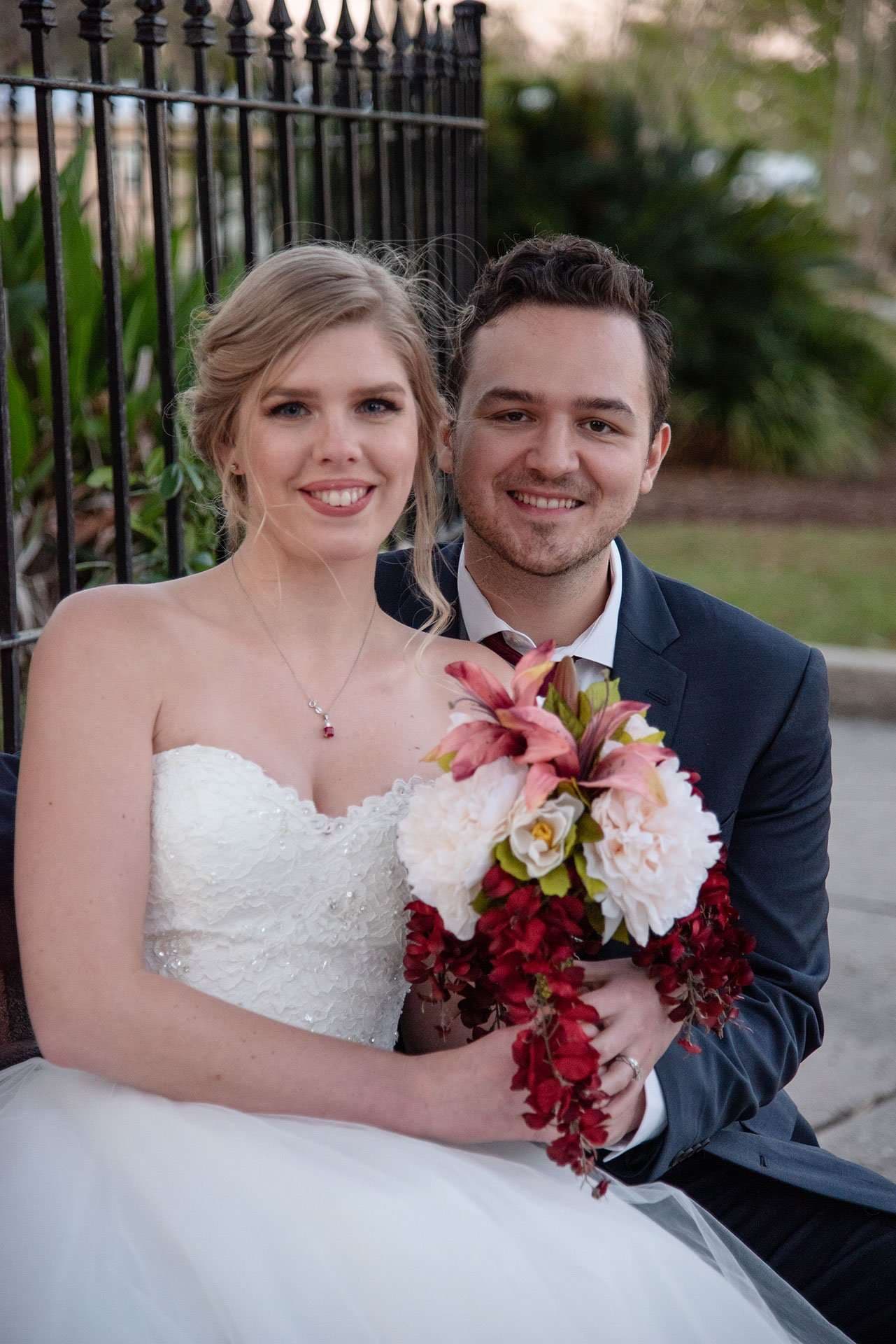 Smiling couple sitting close, bride in strapless white gown holding a red and pink bouquet, groom in navy suit