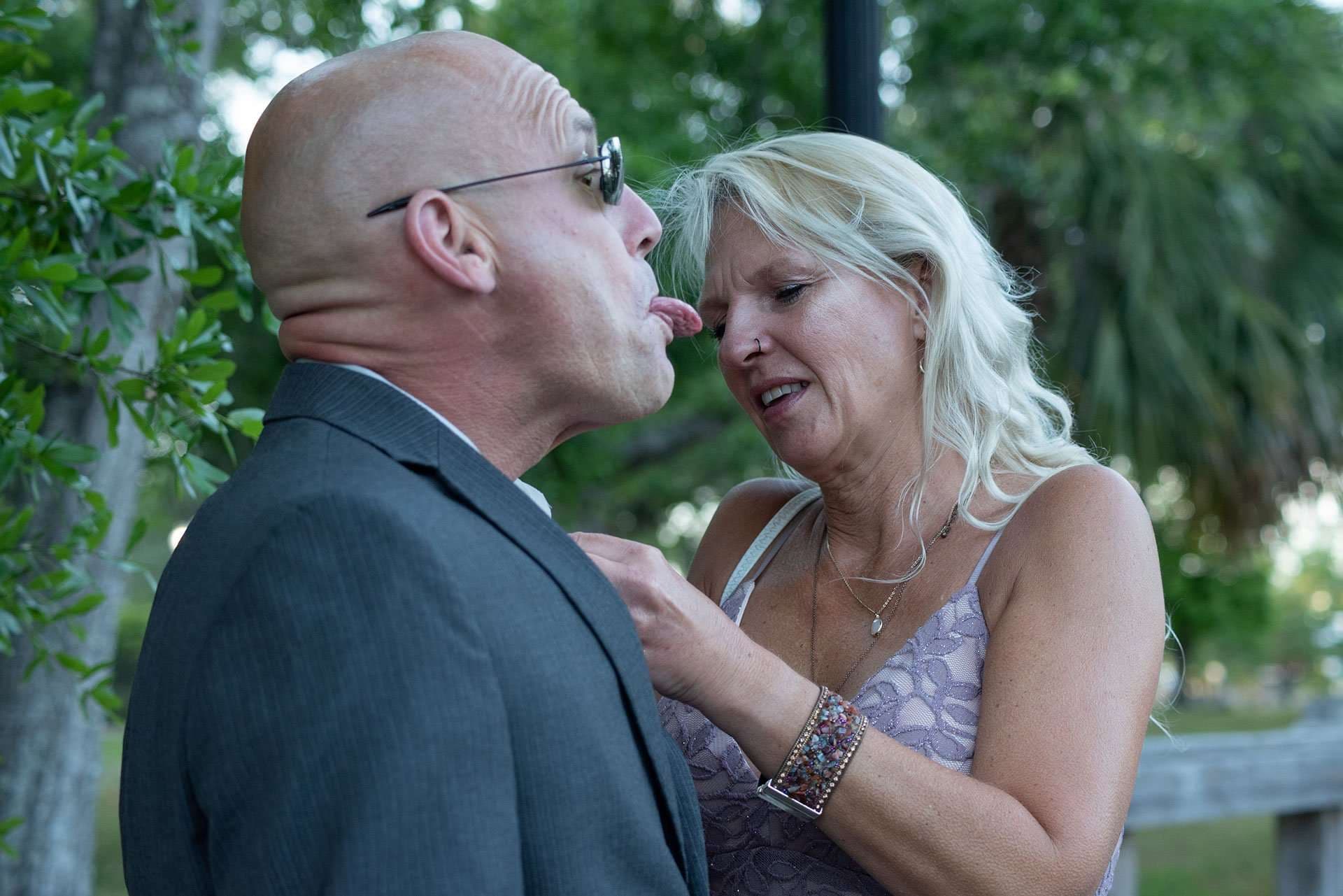 Bald man in glasses and gray suit sticks out his tongue as a blonde woman adjusts his lapel outdoors