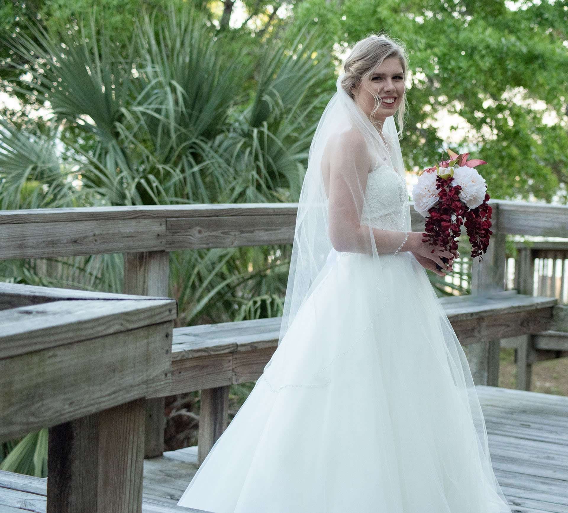 Smiling bride in a white tulle gown and long veil holding a red and white bouquet on a wooden boardwalk