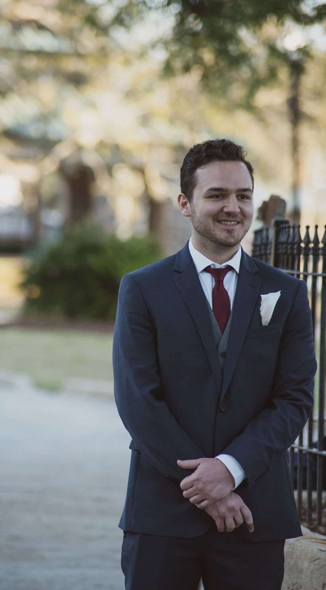 Smiling groom in a navy suit, burgundy tie and white pocket square standing with hands clasped near an iron fence