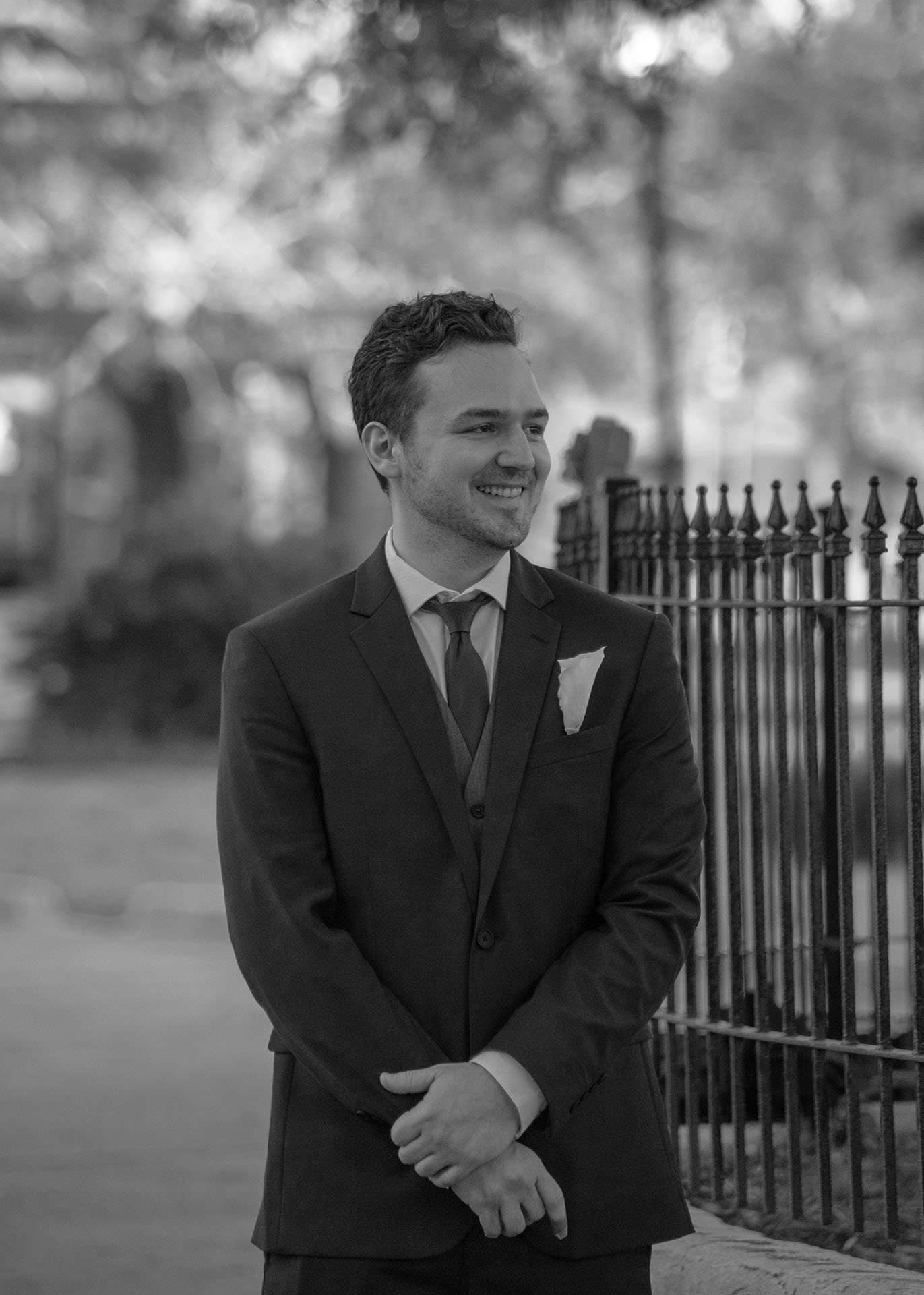 Black-and-white portrait of a smiling man in a suit with pocket square, hands clasped, beside an iron fence