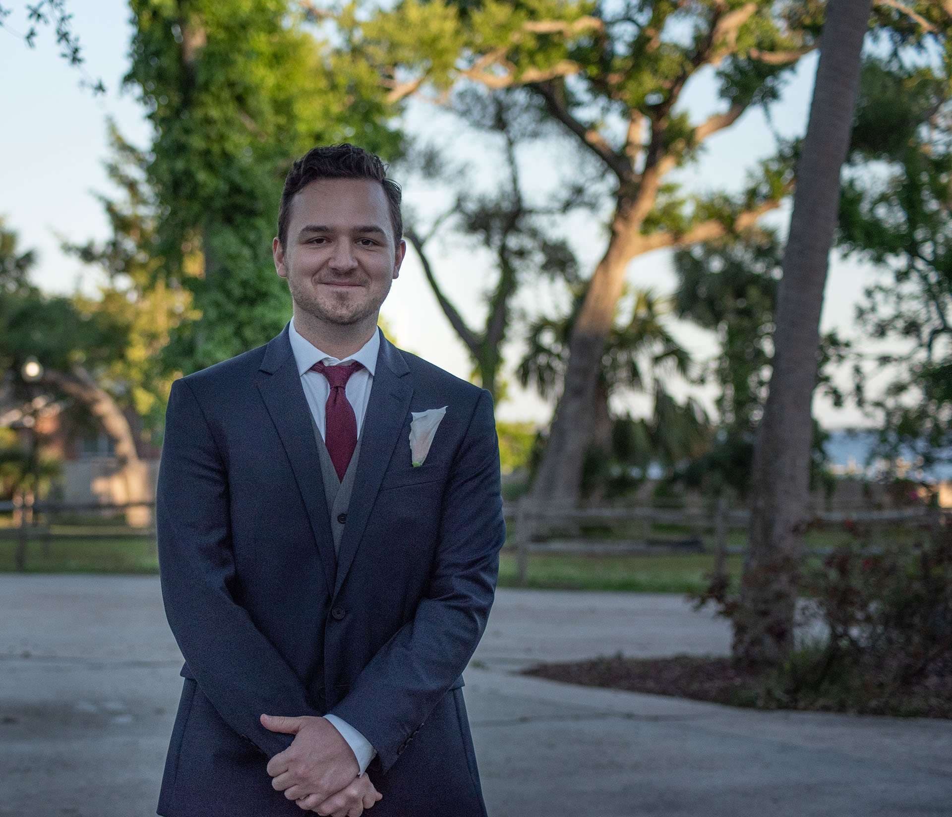 Smiling man in a navy suit, burgundy tie and pocket square standing with hands clasped among trees at dusk
