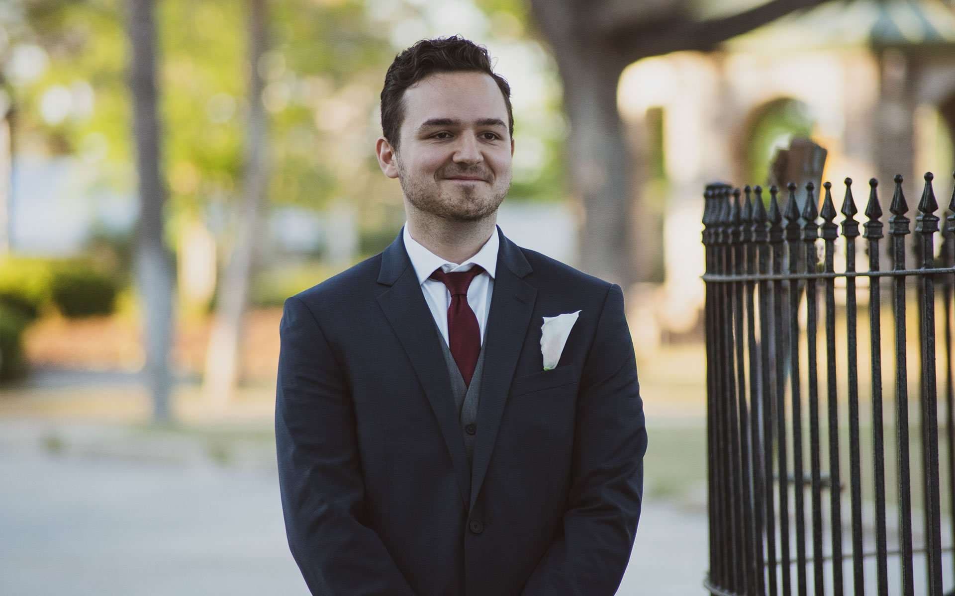 Smiling groom in a navy suit with a burgundy tie and white pocket square stands beside an iron fence