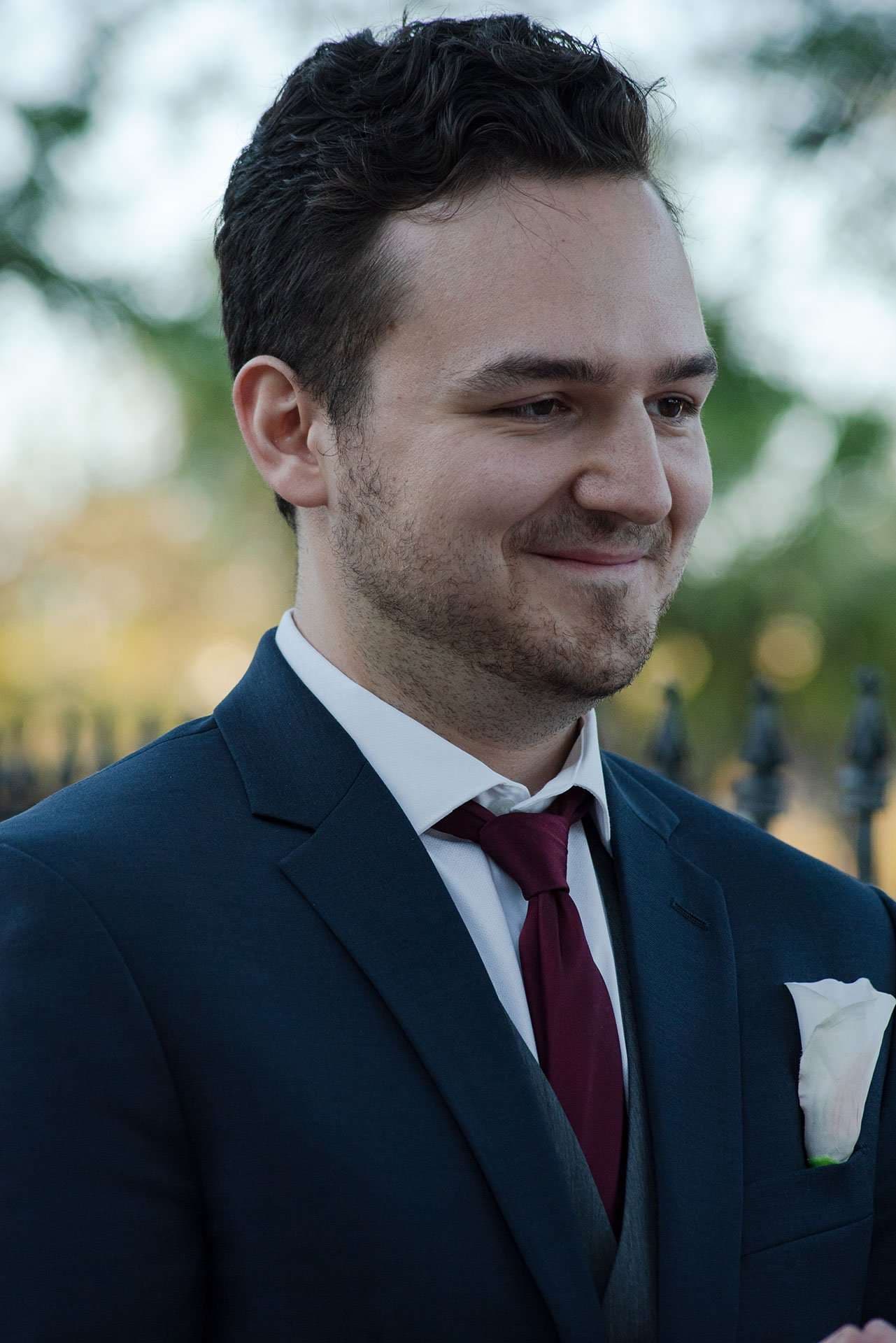 Smiling groom in a navy suit and burgundy tie with a white boutonniere, looking off to the side outdoors