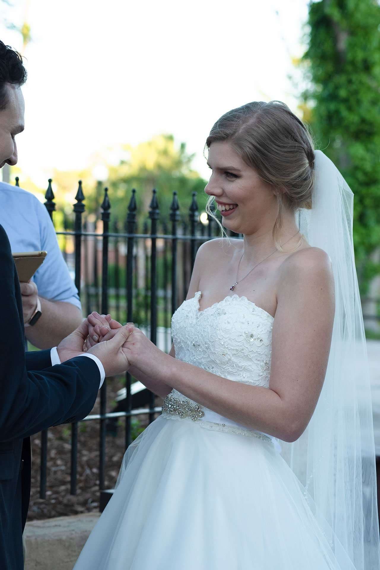 Smiling bride in a strapless white lace gown and veil holds hands with the groom during an outdoor ceremony