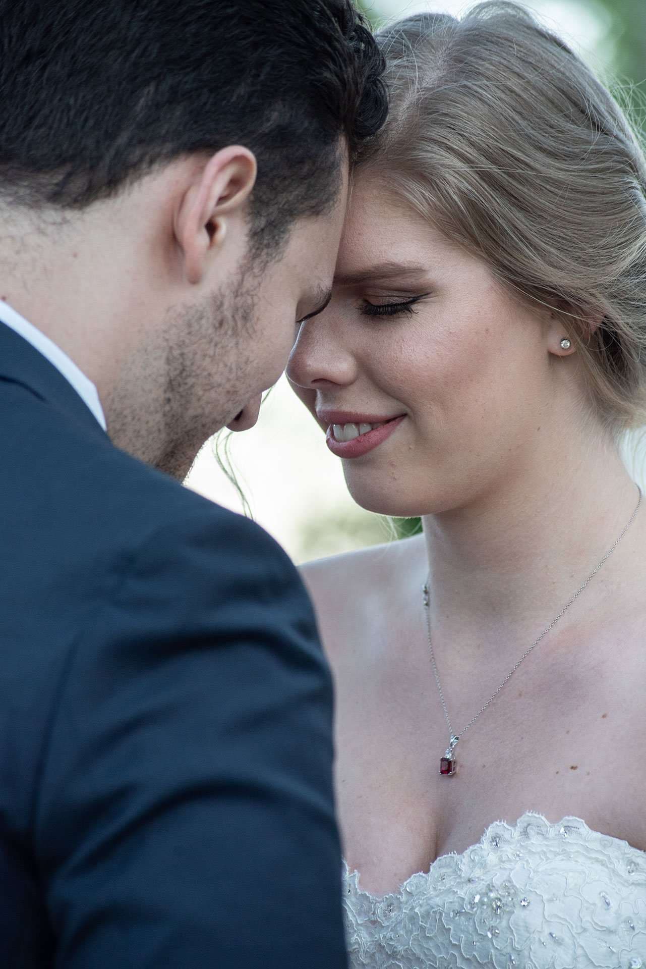 Couple touching foreheads in a tender moment, the bride smiling with closed eyes in a white lace gown and ruby pendant