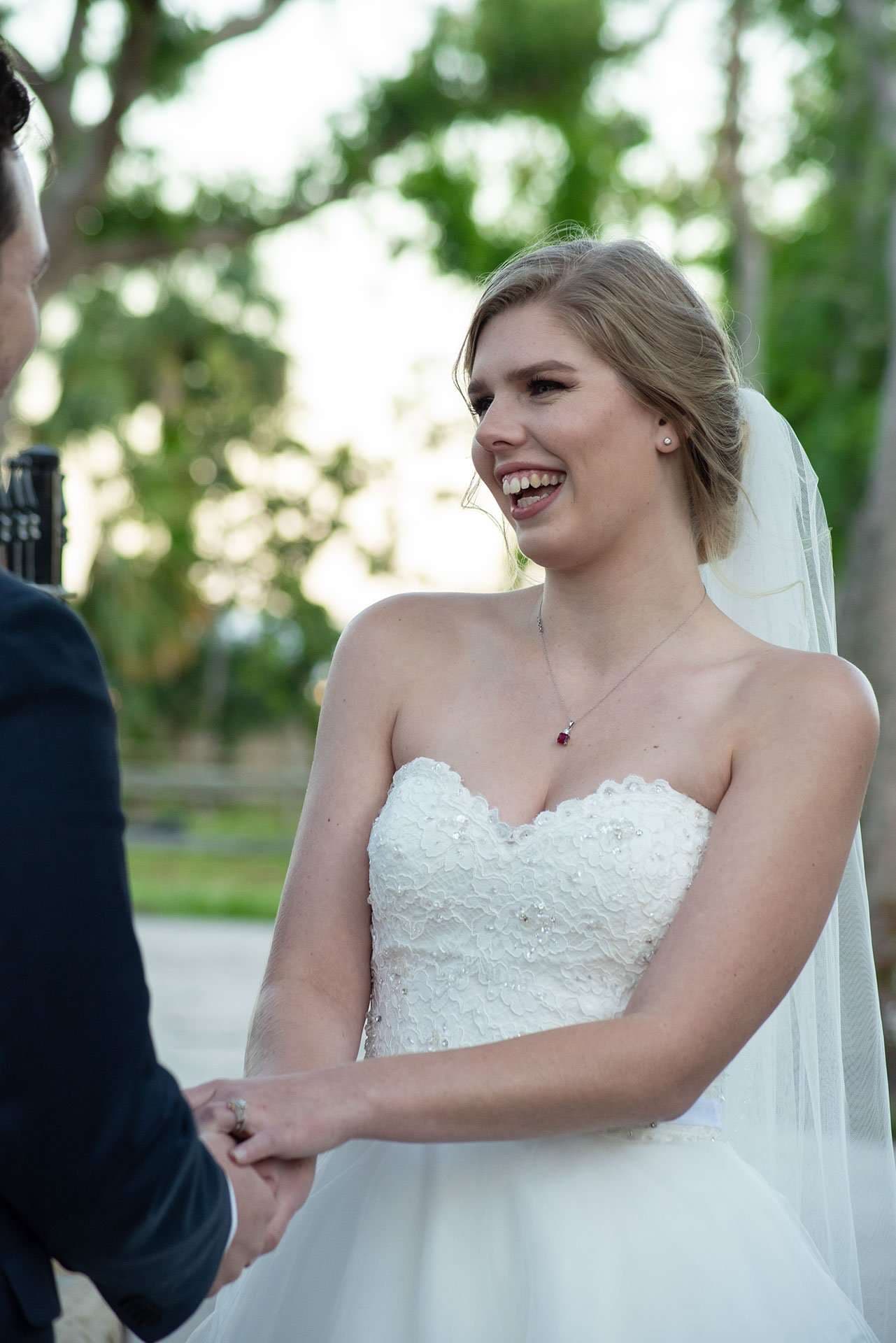 Laughing bride in a strapless white lace gown and veil holding hands with her partner outdoors among trees