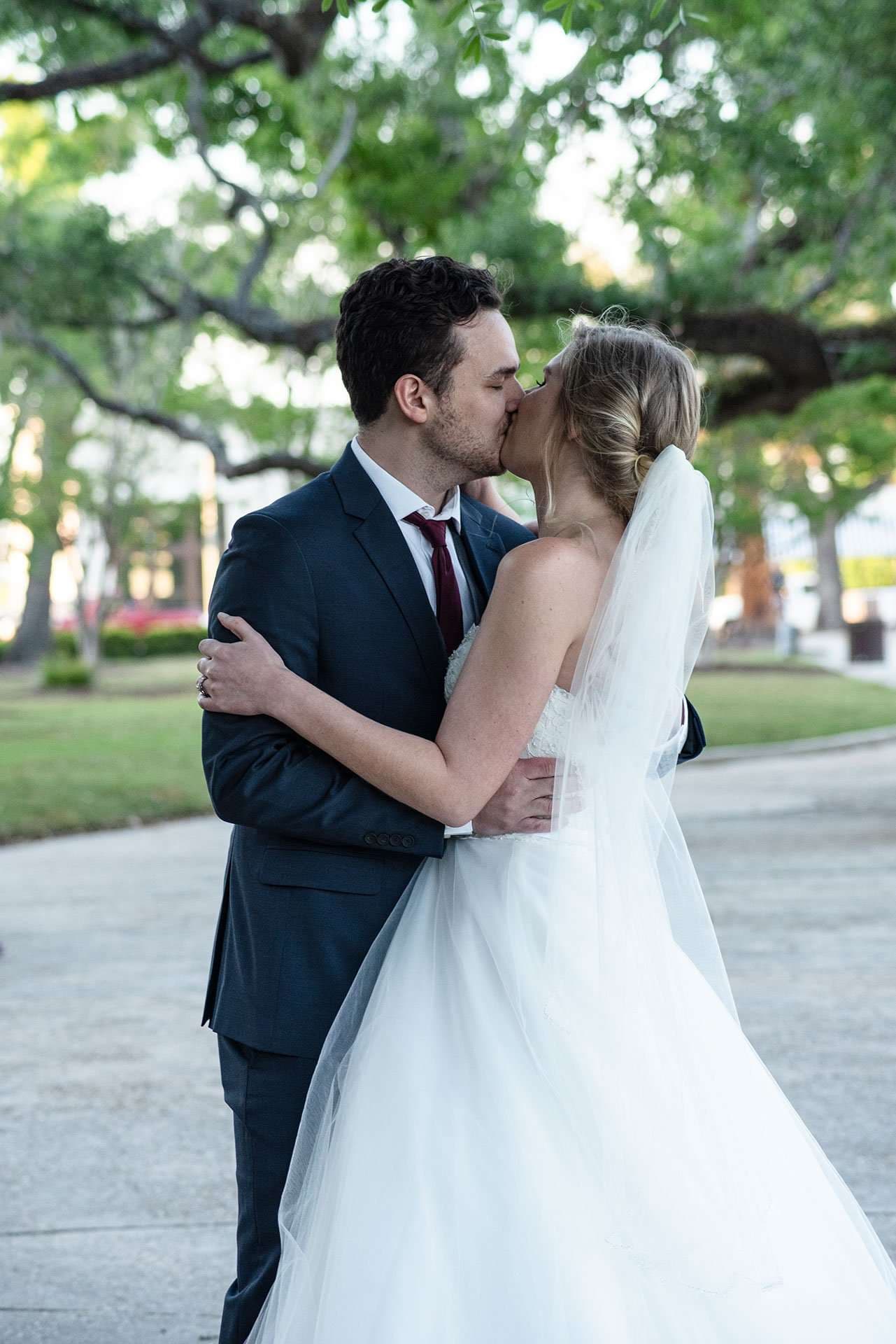 Couple kissing under large oak trees, groom in a navy suit and bride in a white gown with a long veil