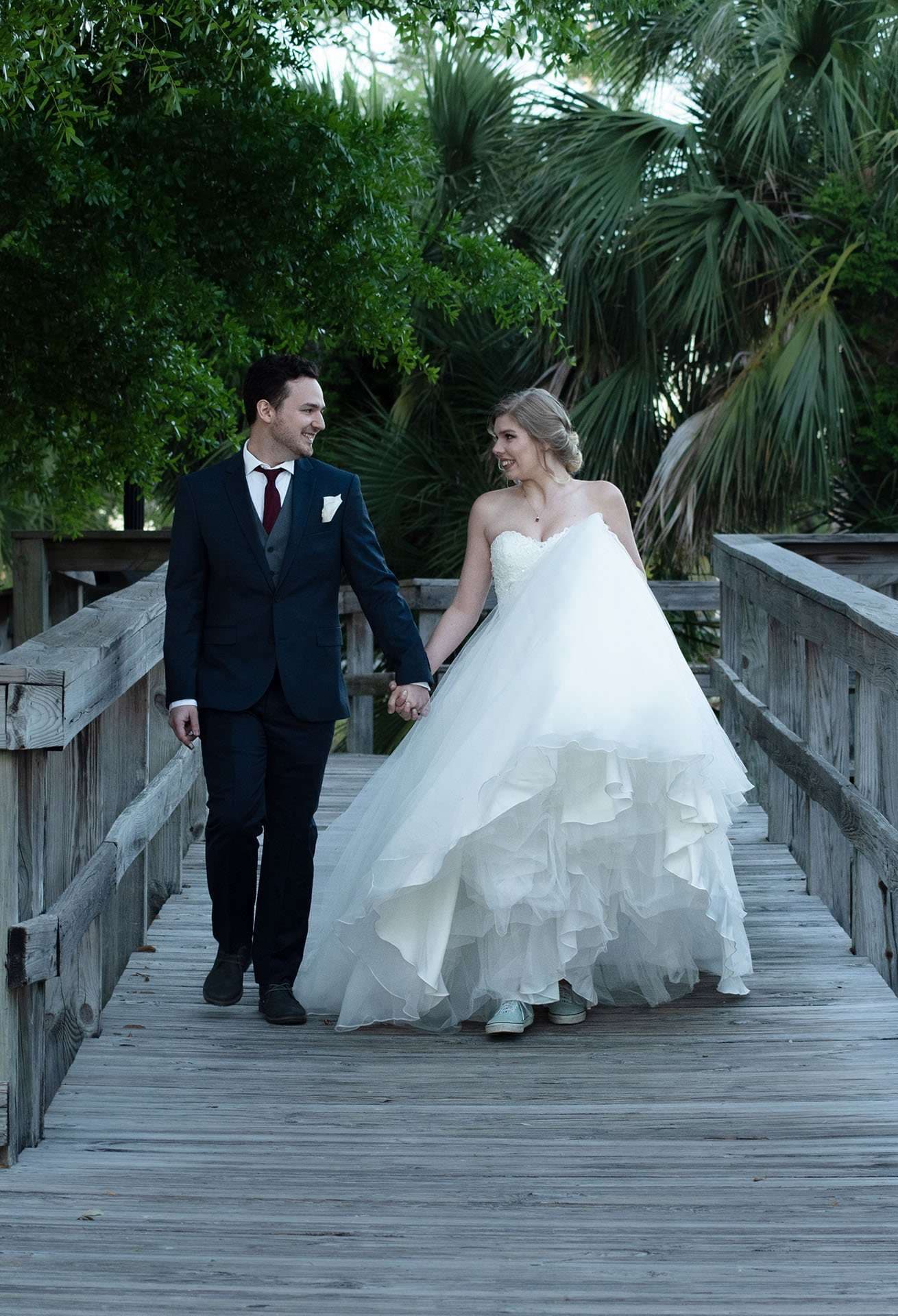 Couple holding hands and smiling while walking along a wooden boardwalk, bride in a full white tulle gown, groom in navy suit