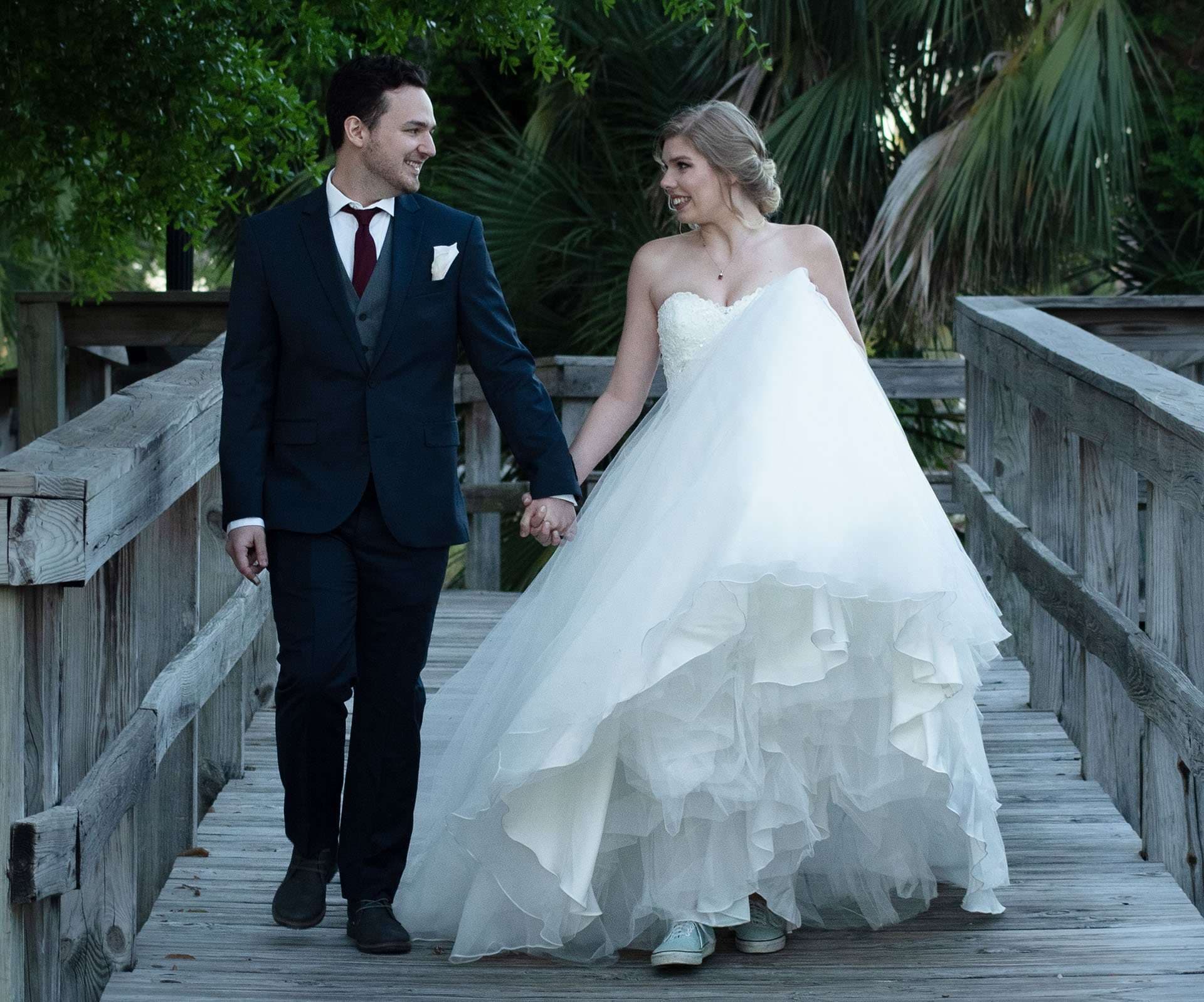 Groom in a navy suit holding hands with his bride in a ruffled white gown, walking on a wooden boardwalk