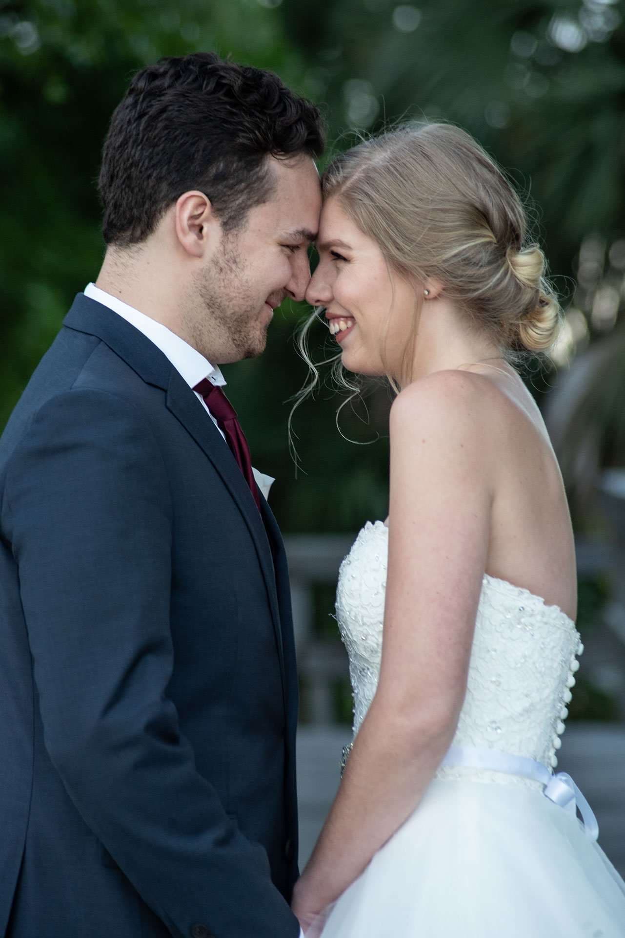 Smiling couple touching foreheads outdoors, bride in a white strapless lace gown, groom in a navy suit and burgundy tie
