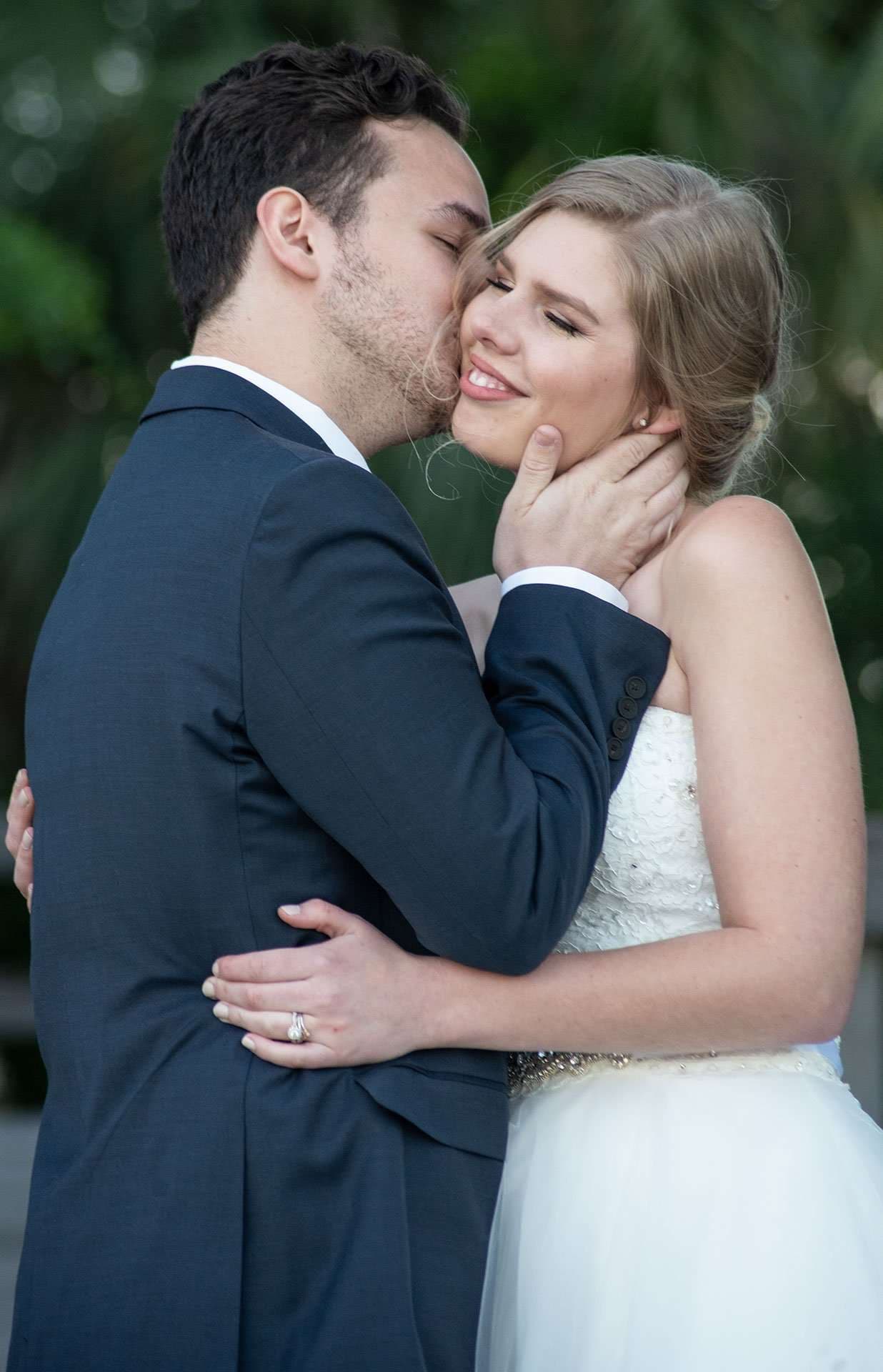 Groom in navy suit kisses smiling bride's cheek, cradling her face, as she wears a white strapless gown outdoors