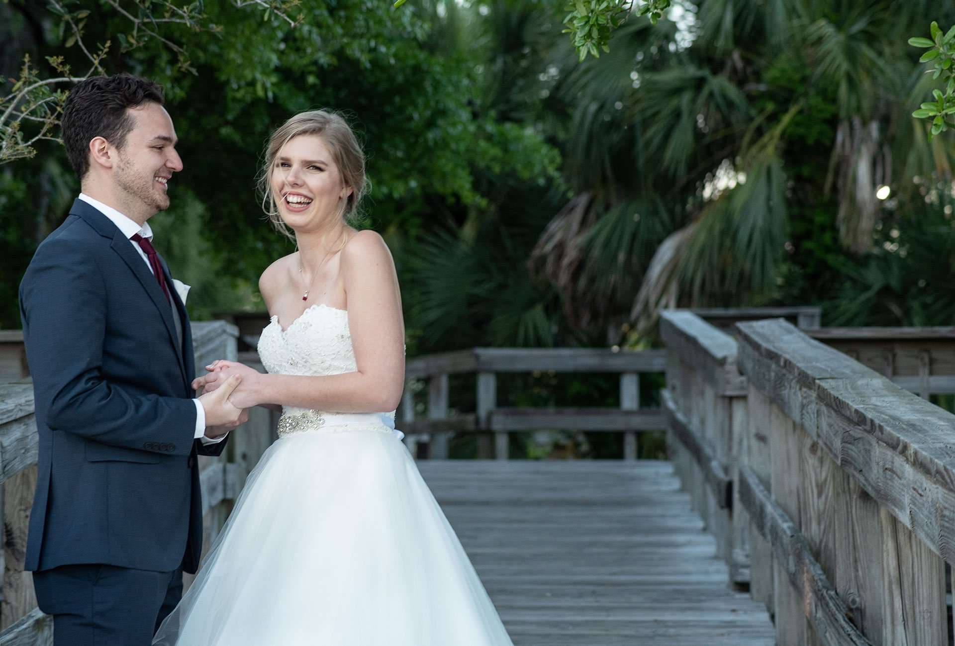 Laughing bride in a white strapless gown holding hands with a groom in a navy suit on a wooden boardwalk