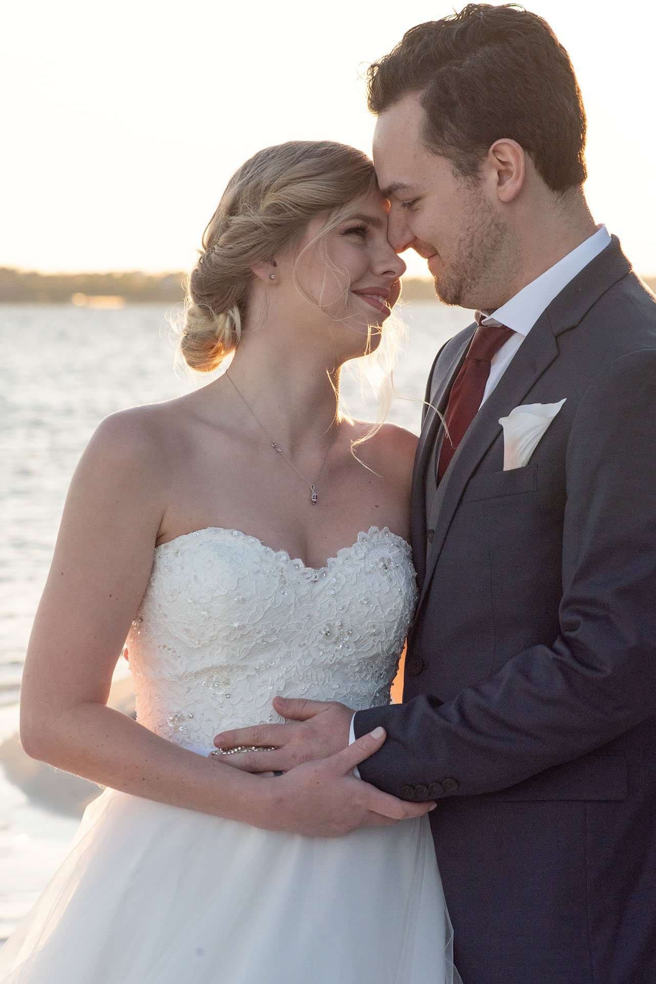 Bride in a strapless lace gown and groom in a gray suit touch foreheads and smile by the water at sunset
