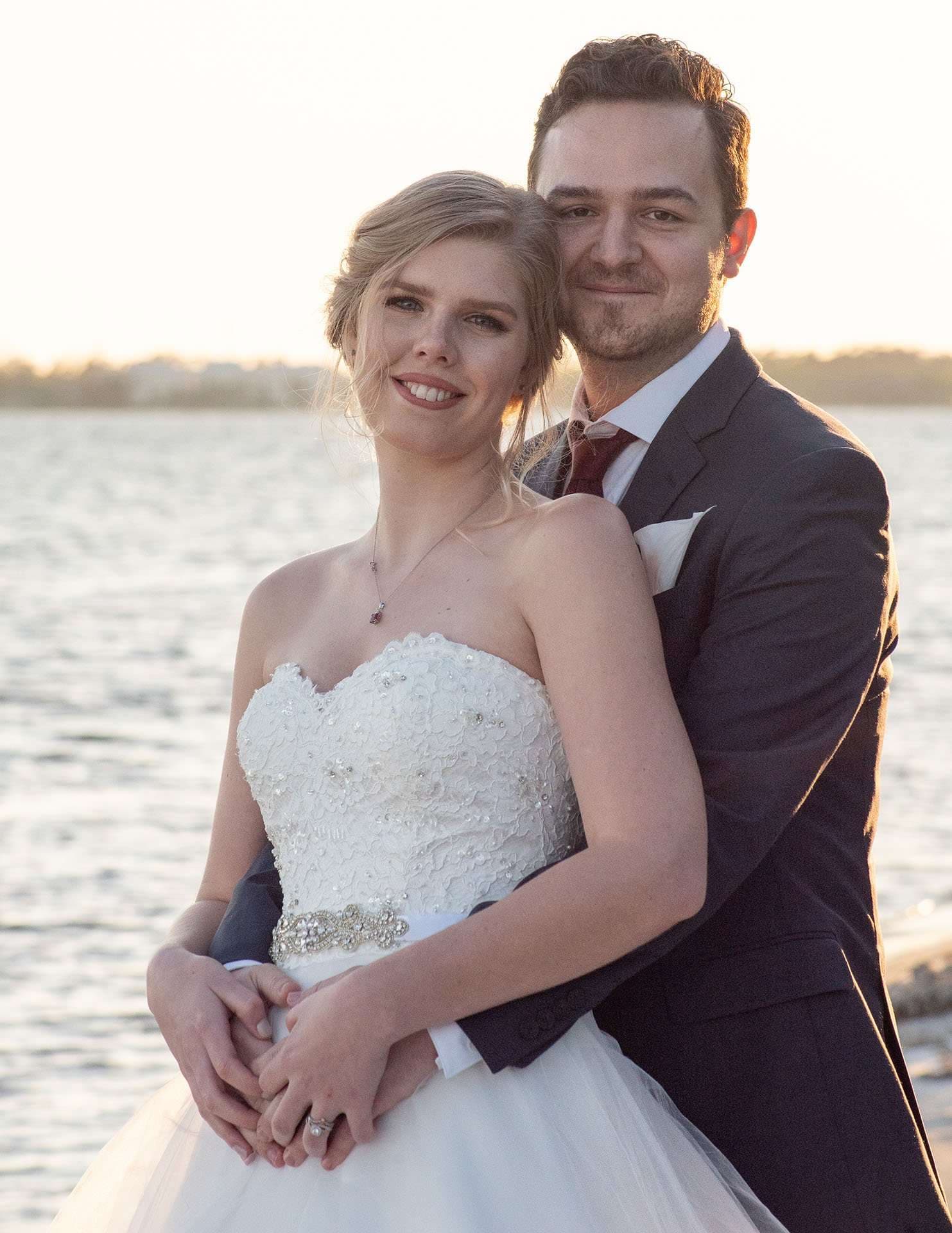 Groom embracing his smiling bride from behind beside the water at sunset, she wears a beaded white strapless gown