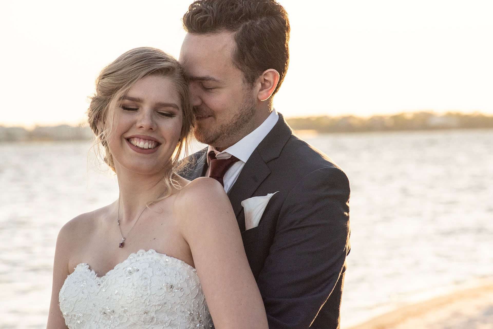 Smiling bride in a strapless beaded gown embraced from behind by her groom in a navy suit beside the water at sunset