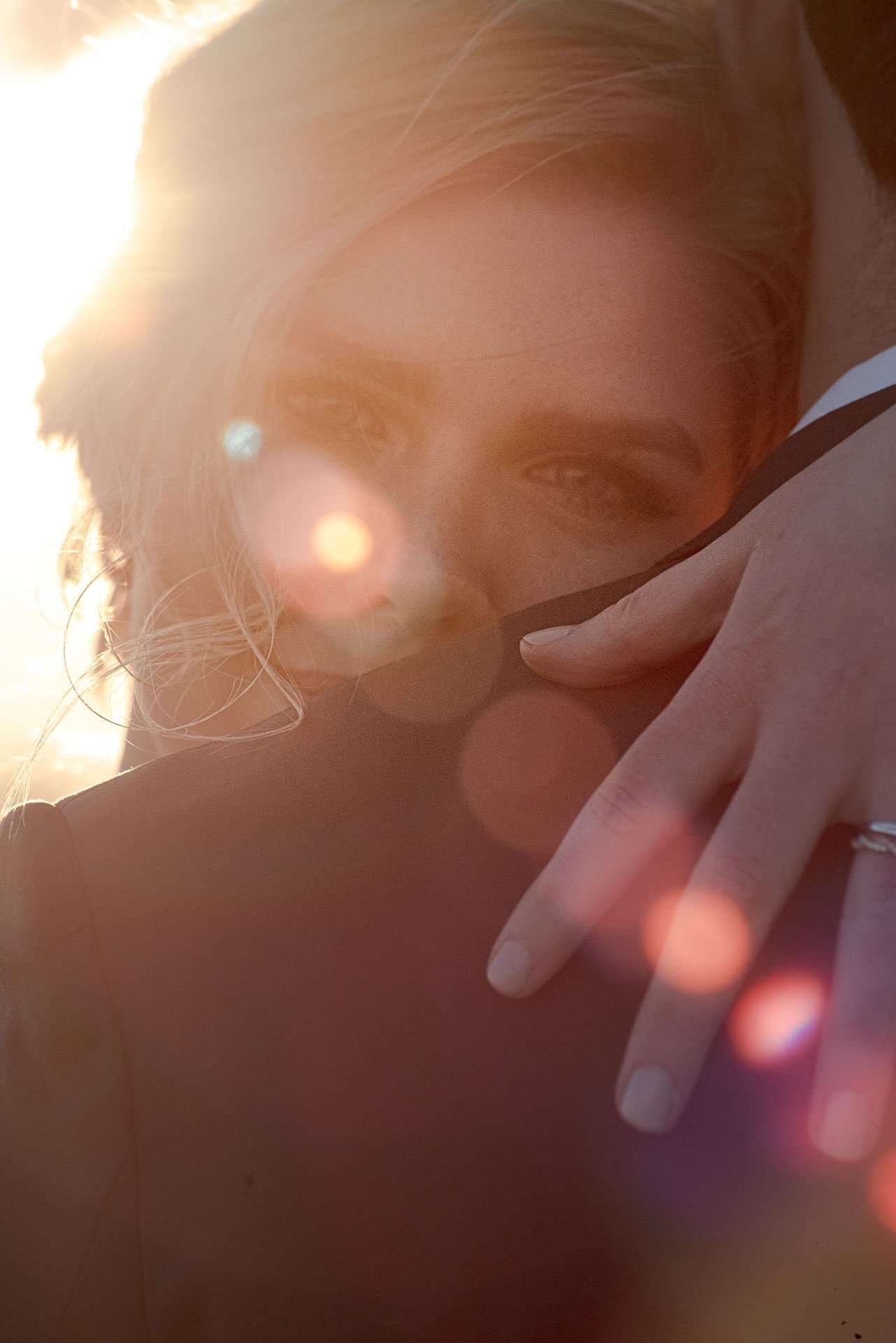 Close-up of a woman's face half hidden by a dark jacket collar, a ringed hand on her shoulder, in golden backlight