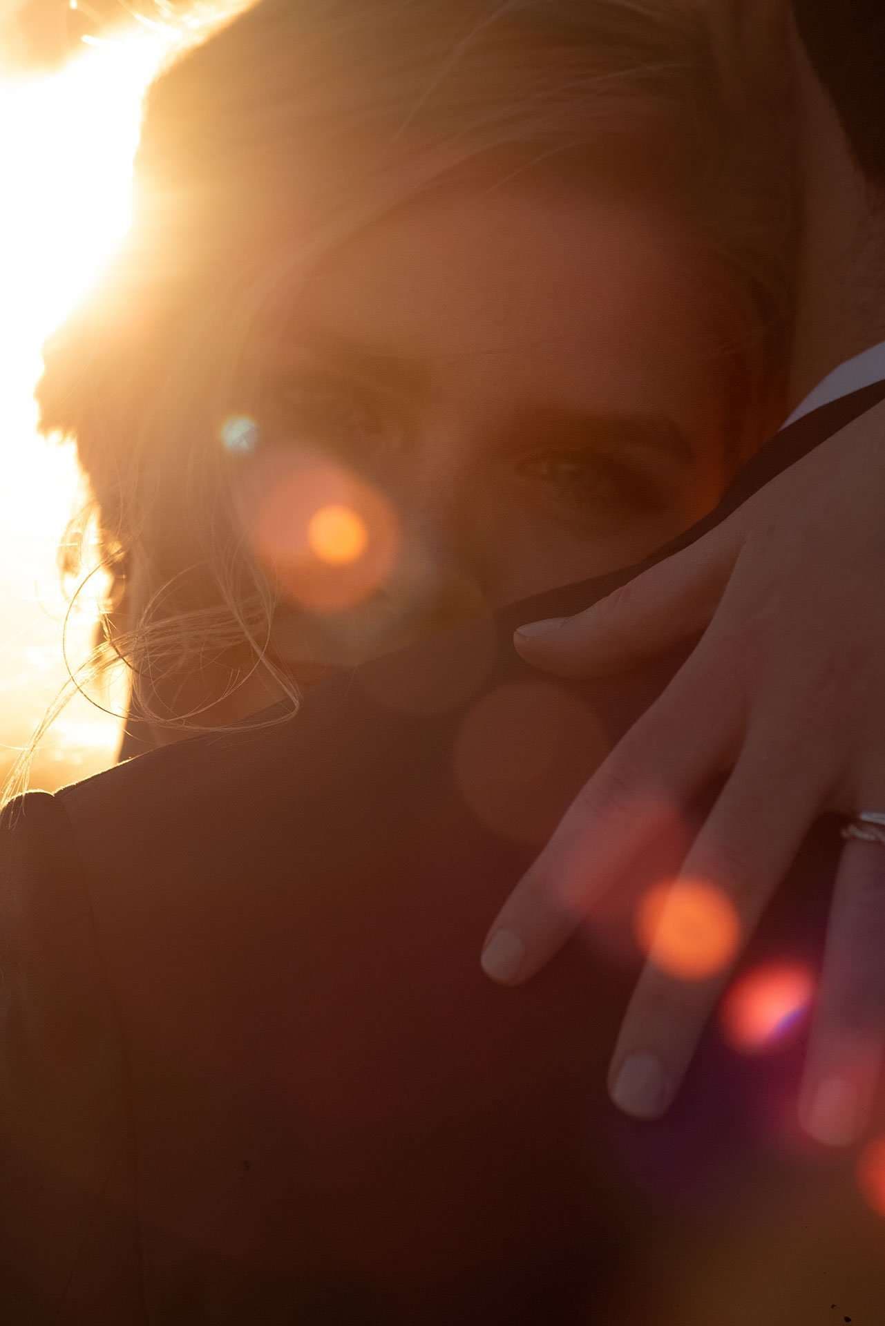 Close-up of a woman's face at sunset with a hand wearing a ring resting on her shoulder, warm lens flare