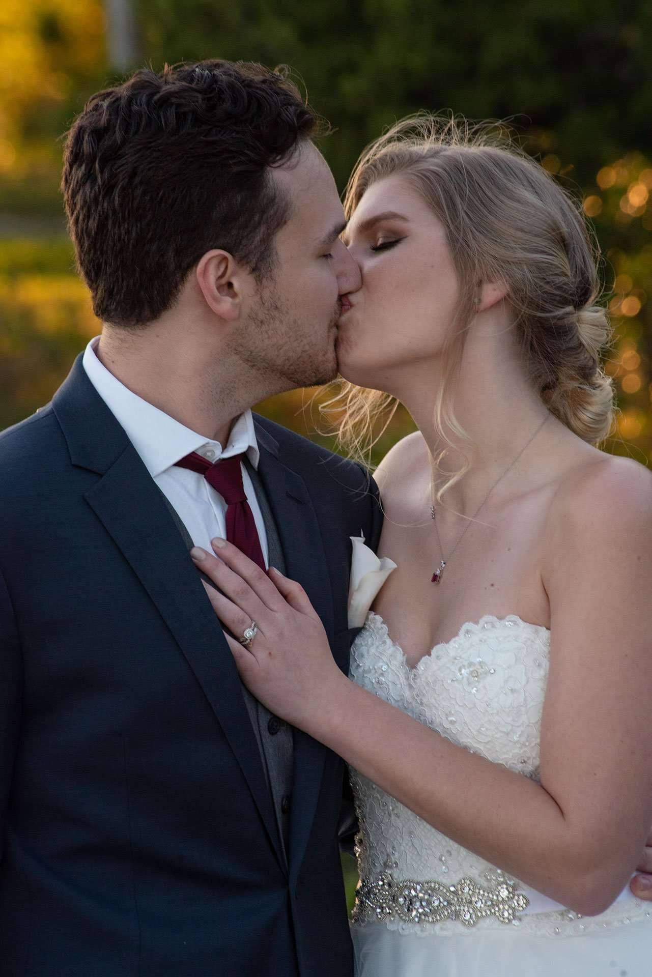 Couple kissing outdoors at golden hour, bride in a strapless beaded white gown and groom in a navy suit with burgundy tie