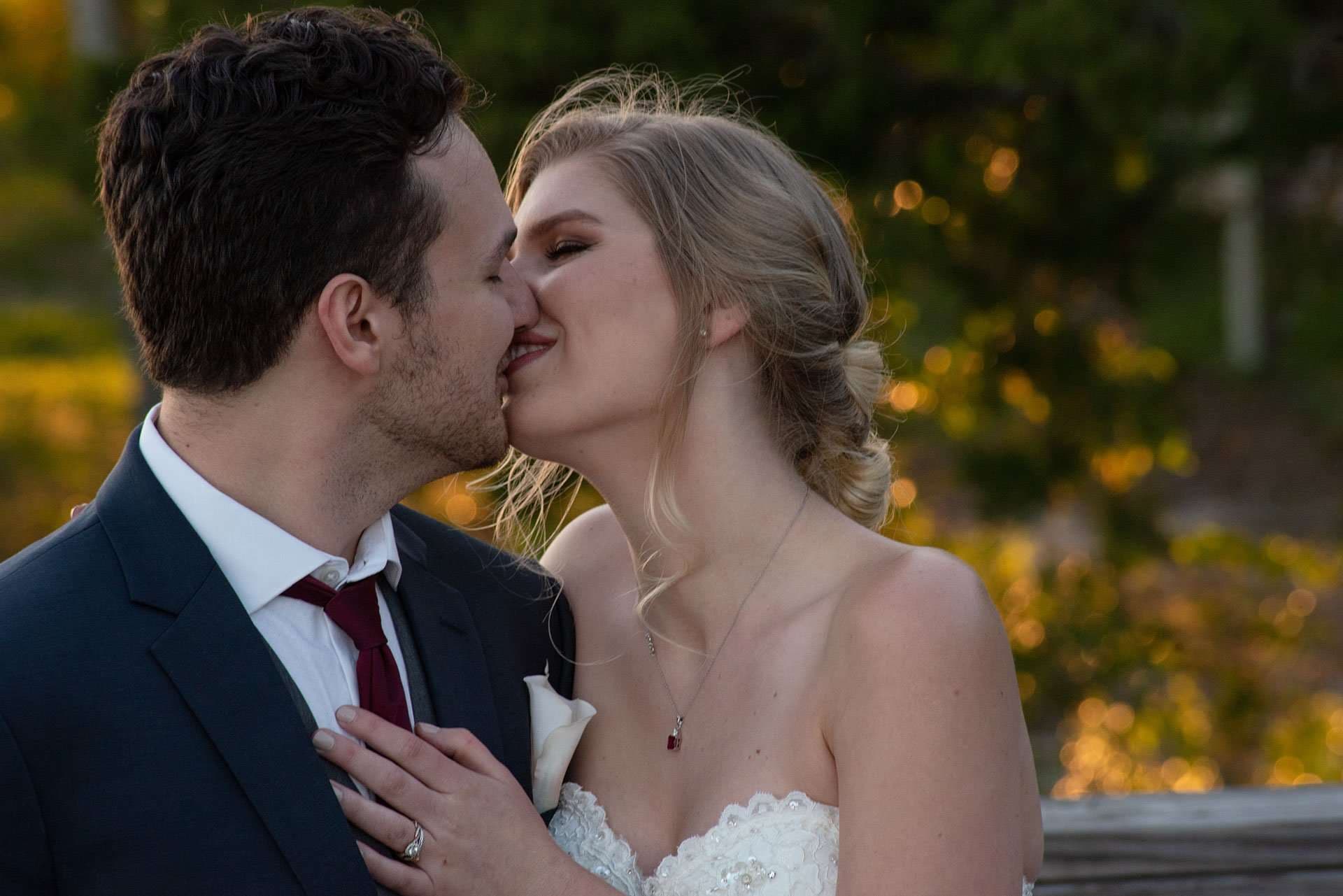 Couple kissing outdoors at golden hour, groom in navy suit with burgundy tie, bride in strapless lace gown