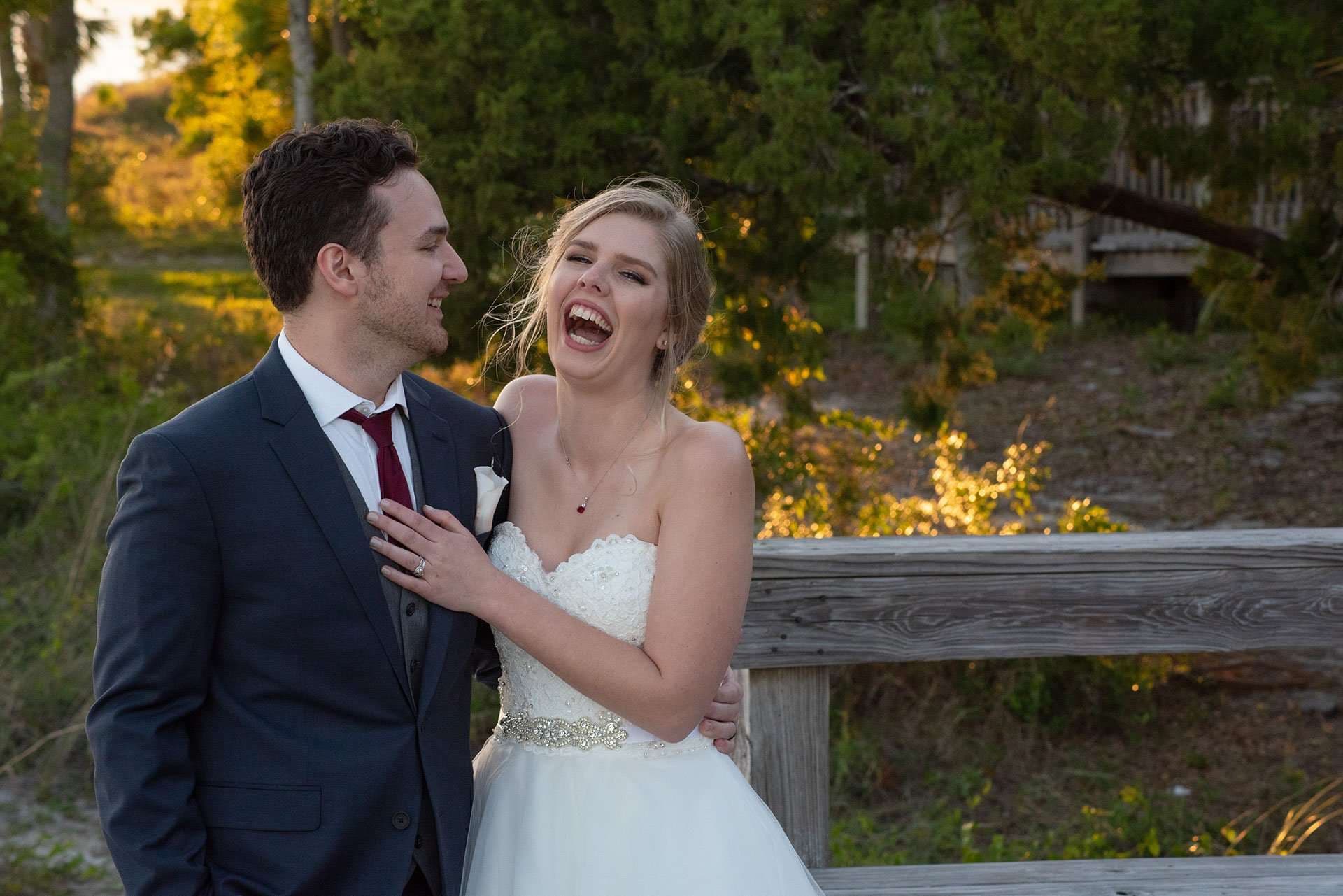 Bride in a strapless white gown laughing joyfully as she embraces her smiling groom in a navy suit by a wooden railing