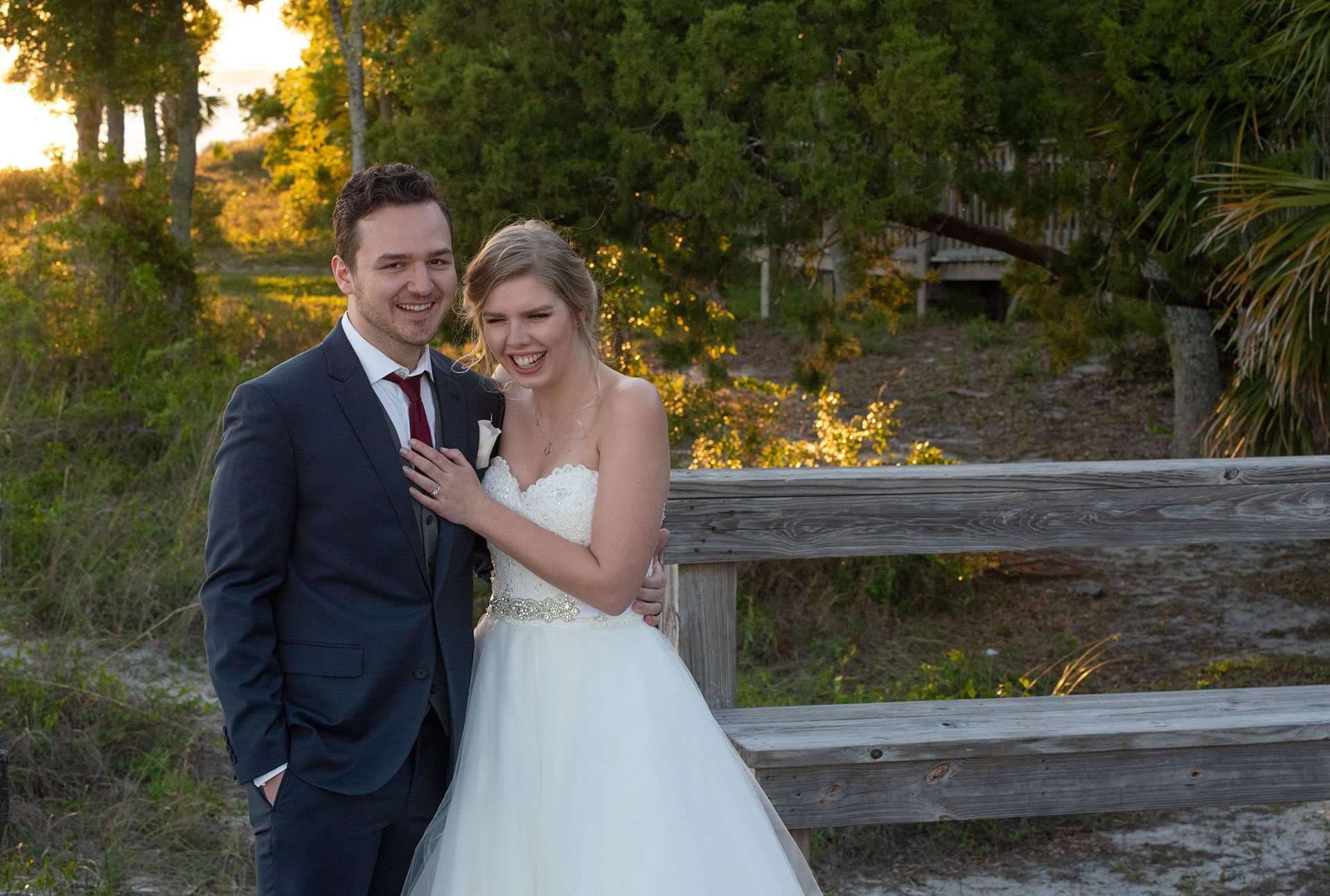 Smiling bride in a white strapless gown and groom in a navy suit embracing beside a wooden railing at sunset