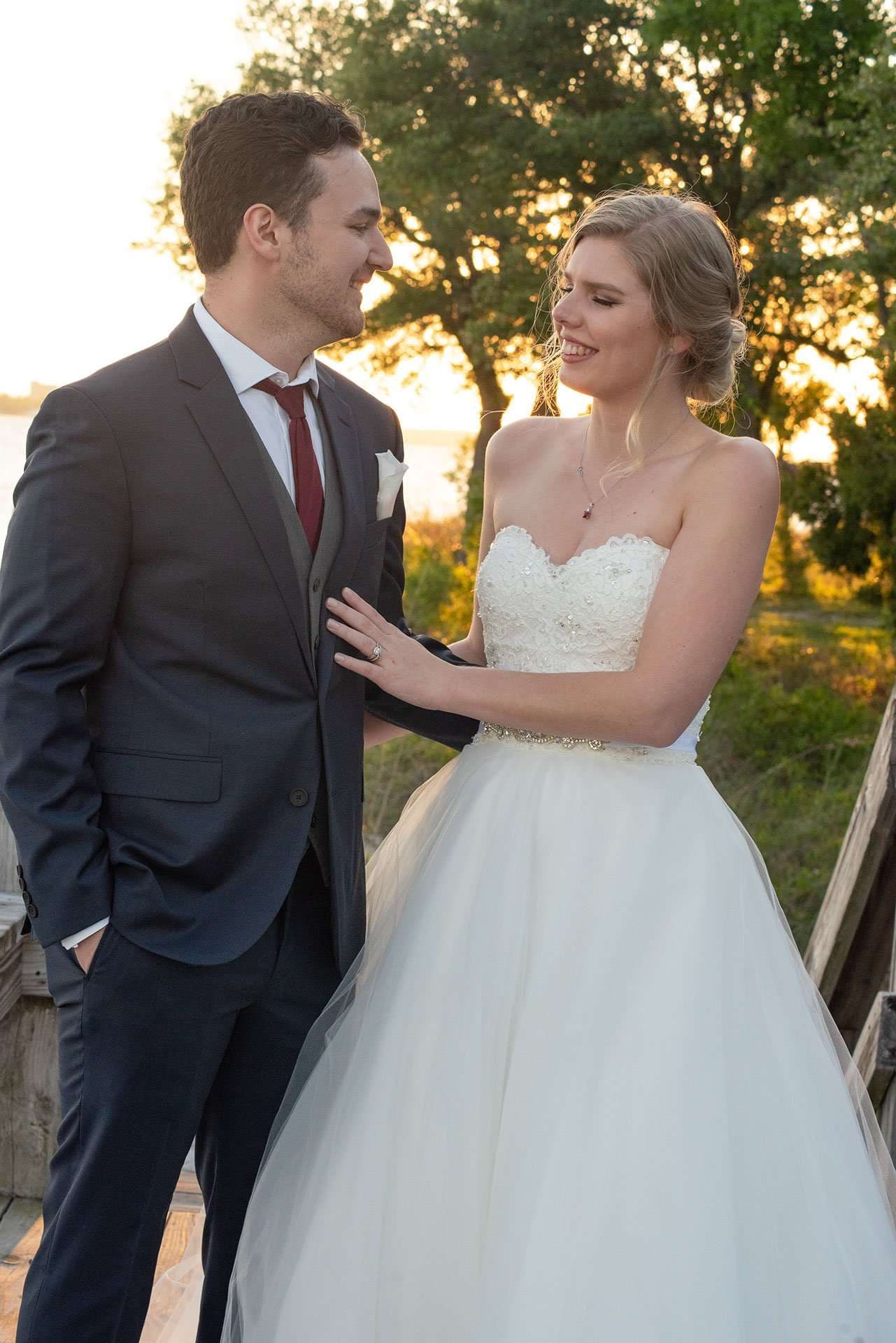 Smiling bride in a strapless white gown touches her groom's chest as they stand by the water at sunset