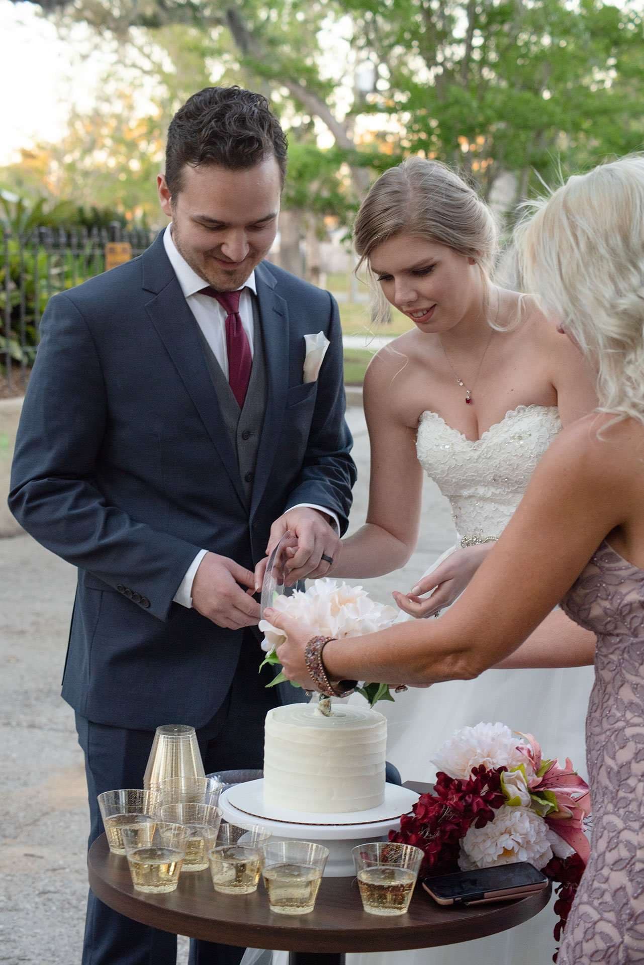 Couple in navy suit and white gown cutting a small white cake as a guest helps, with champagne glasses nearby