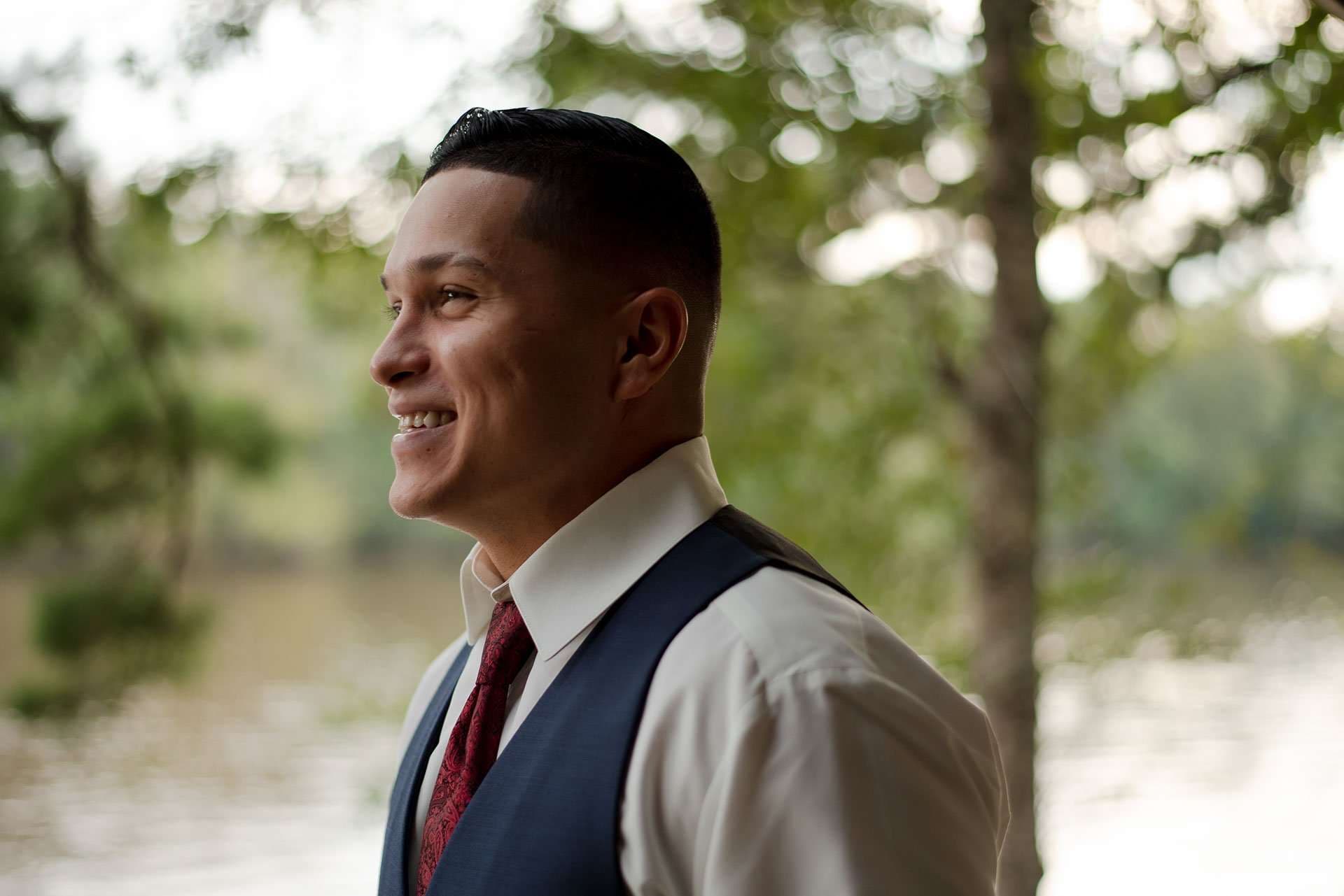 Smiling man in profile wearing a navy vest and burgundy tie, with sunlit trees and water blurred behind him