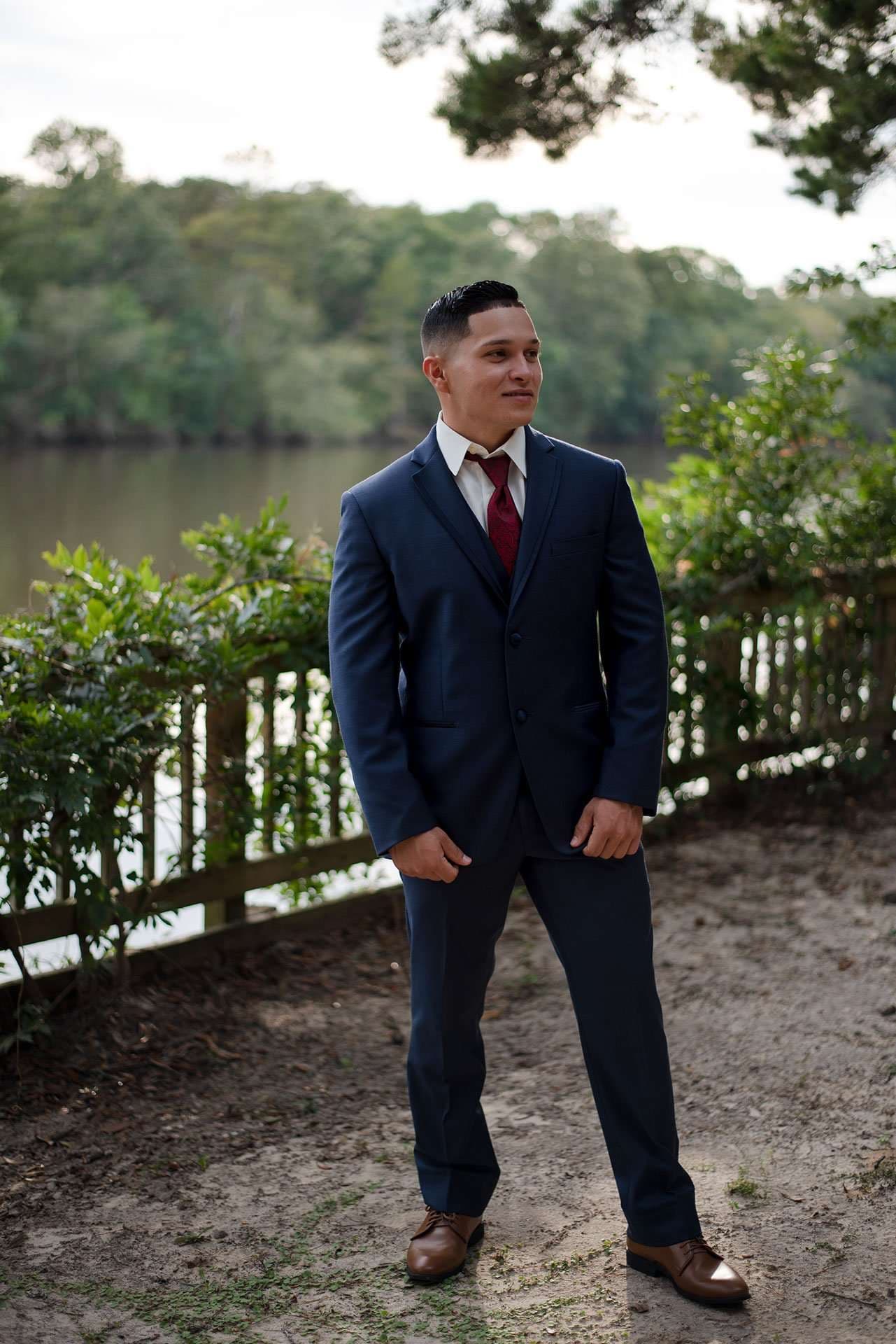 Man in a navy suit and burgundy tie standing on a sandy path by a wooden fence and calm river