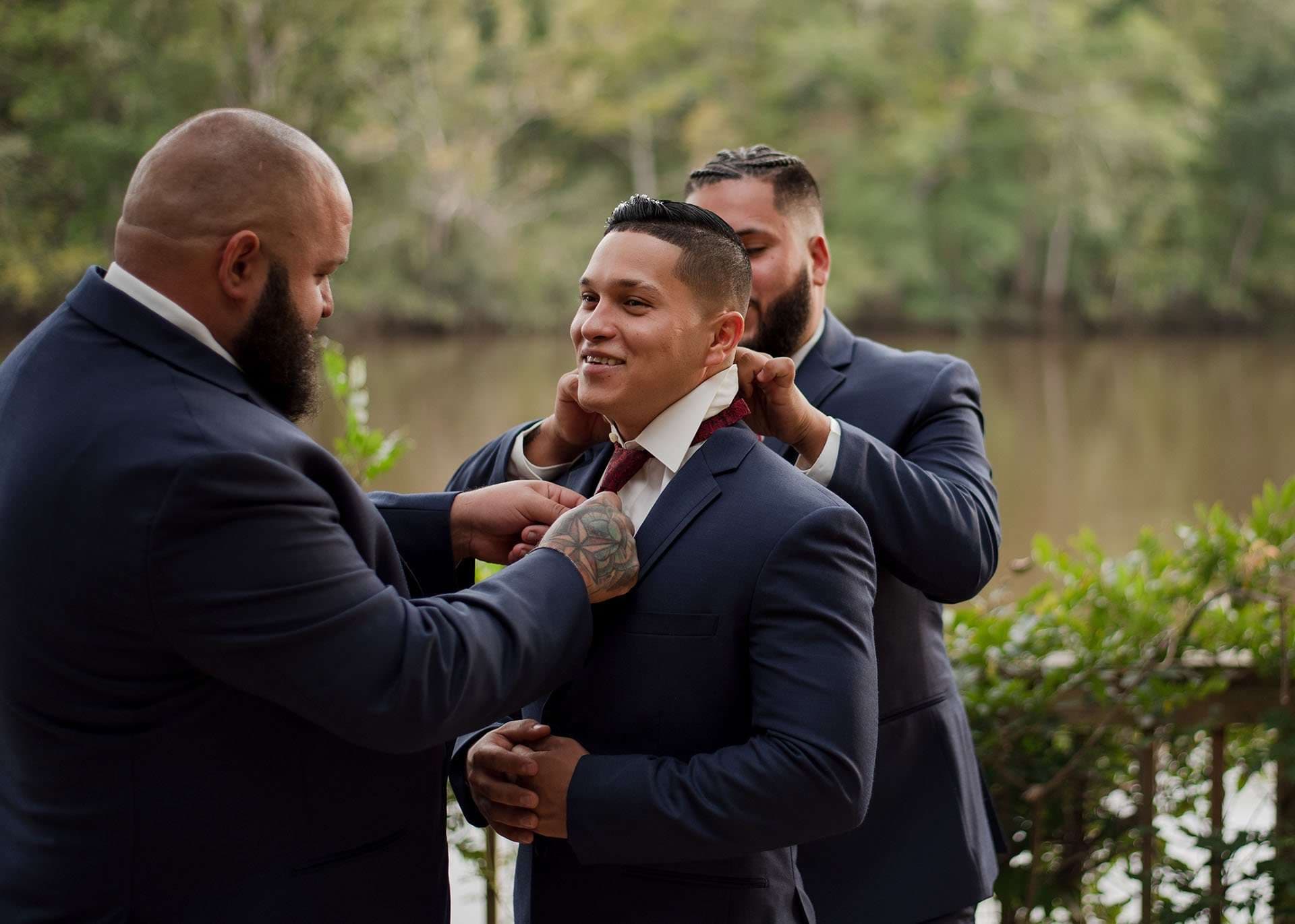Two men in navy suits adjust the burgundy tie and collar of a smiling groom beside a lake