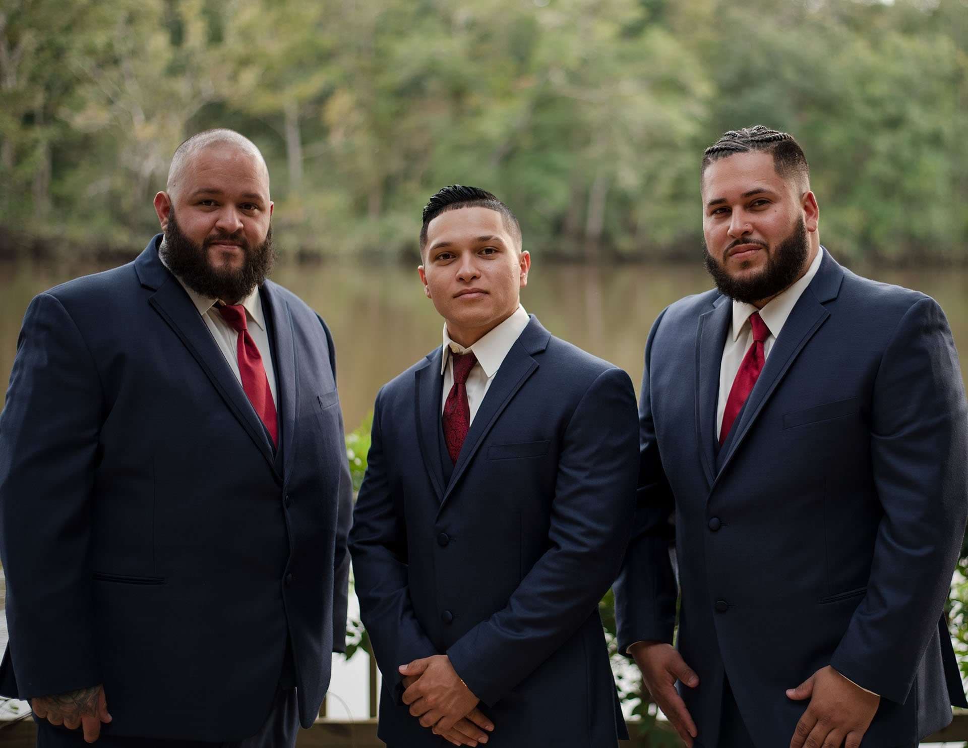 Three men in navy suits and red ties standing together in front of a calm lake and trees