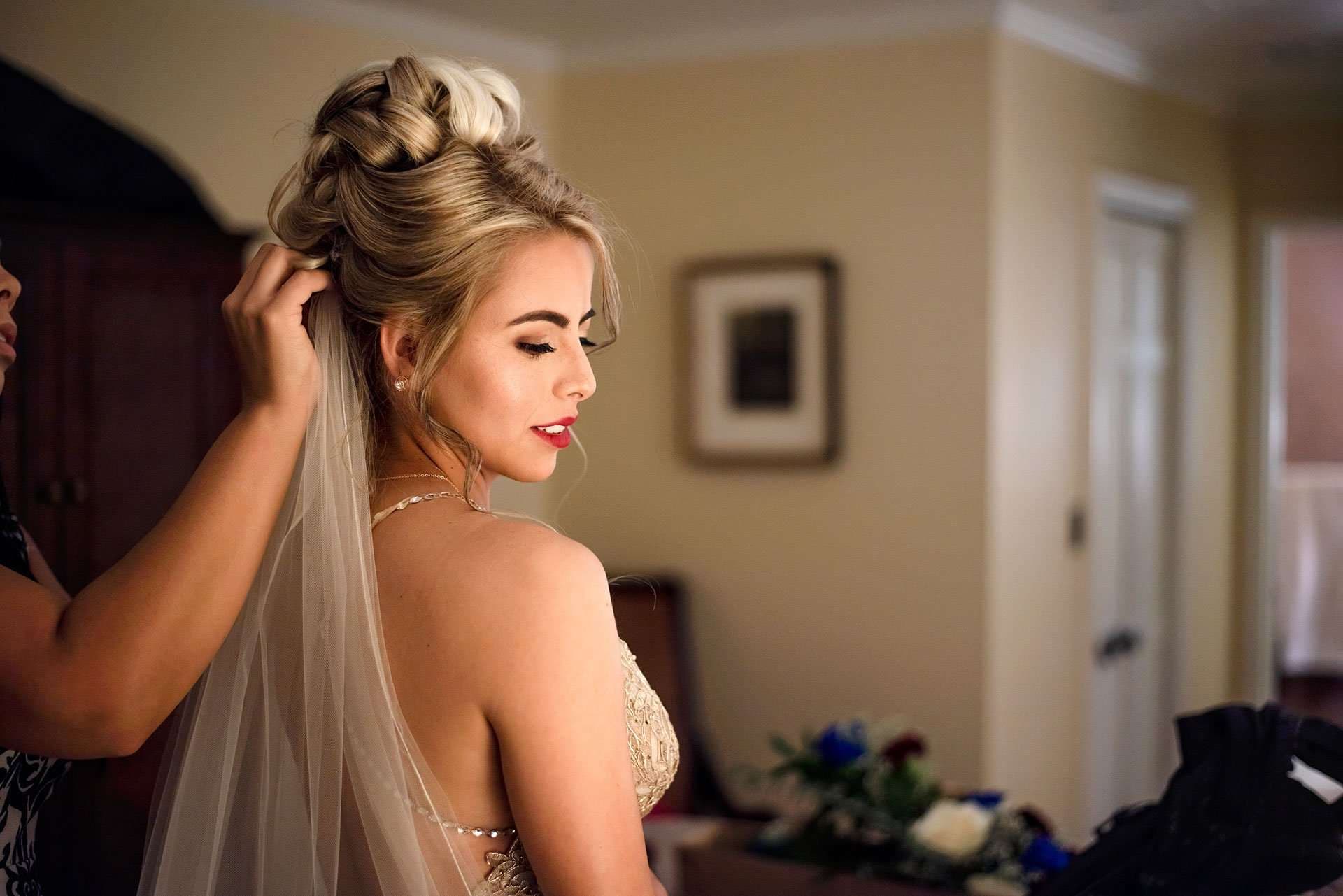 Bride in a beaded gown with an updo has her veil pinned in place by a helper while getting ready