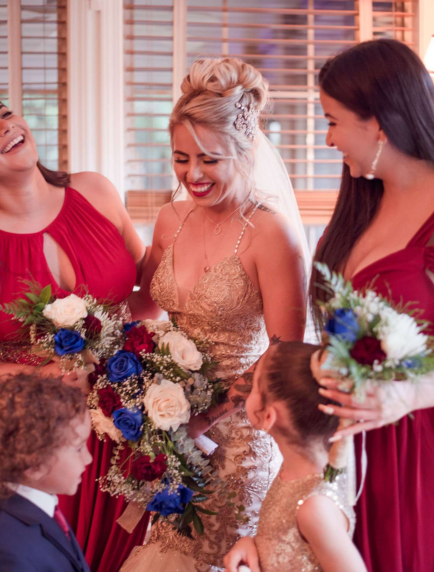 Smiling bride in gold lace gown laughing with bridesmaids in red holding blue and red bouquets, a flower girl beside her
