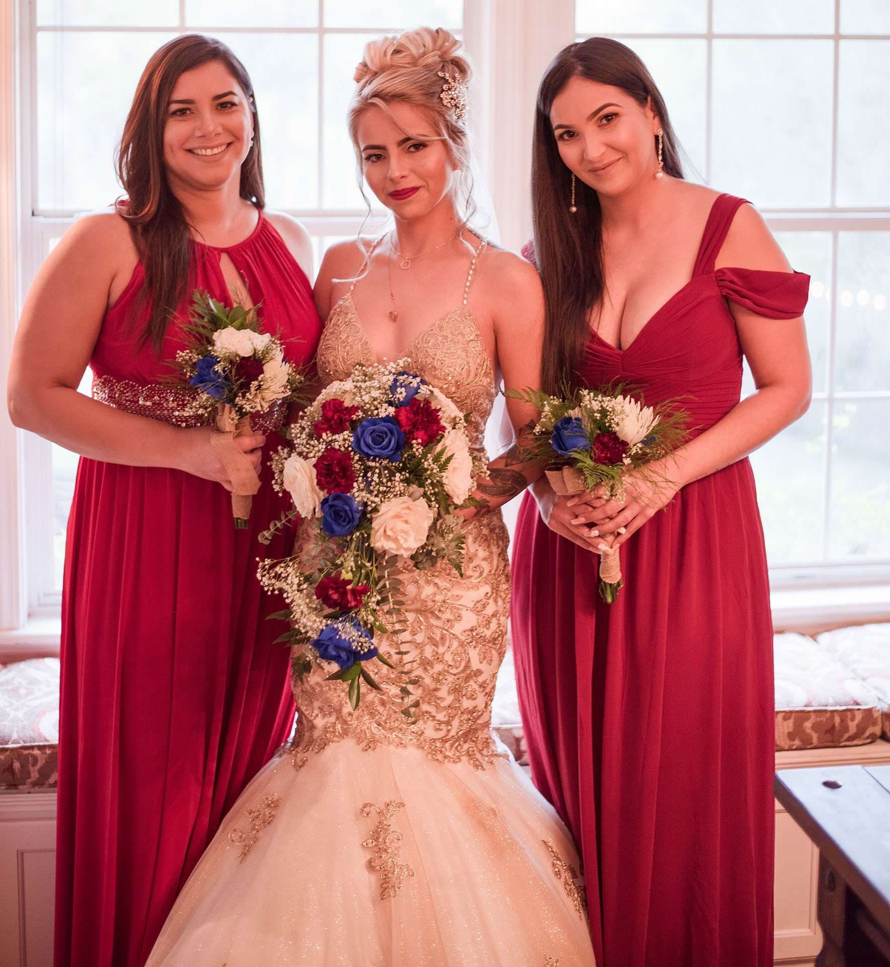 Bride in a gold-embroidered mermaid gown holding a blue and red bouquet, flanked by two bridesmaids in red dresses