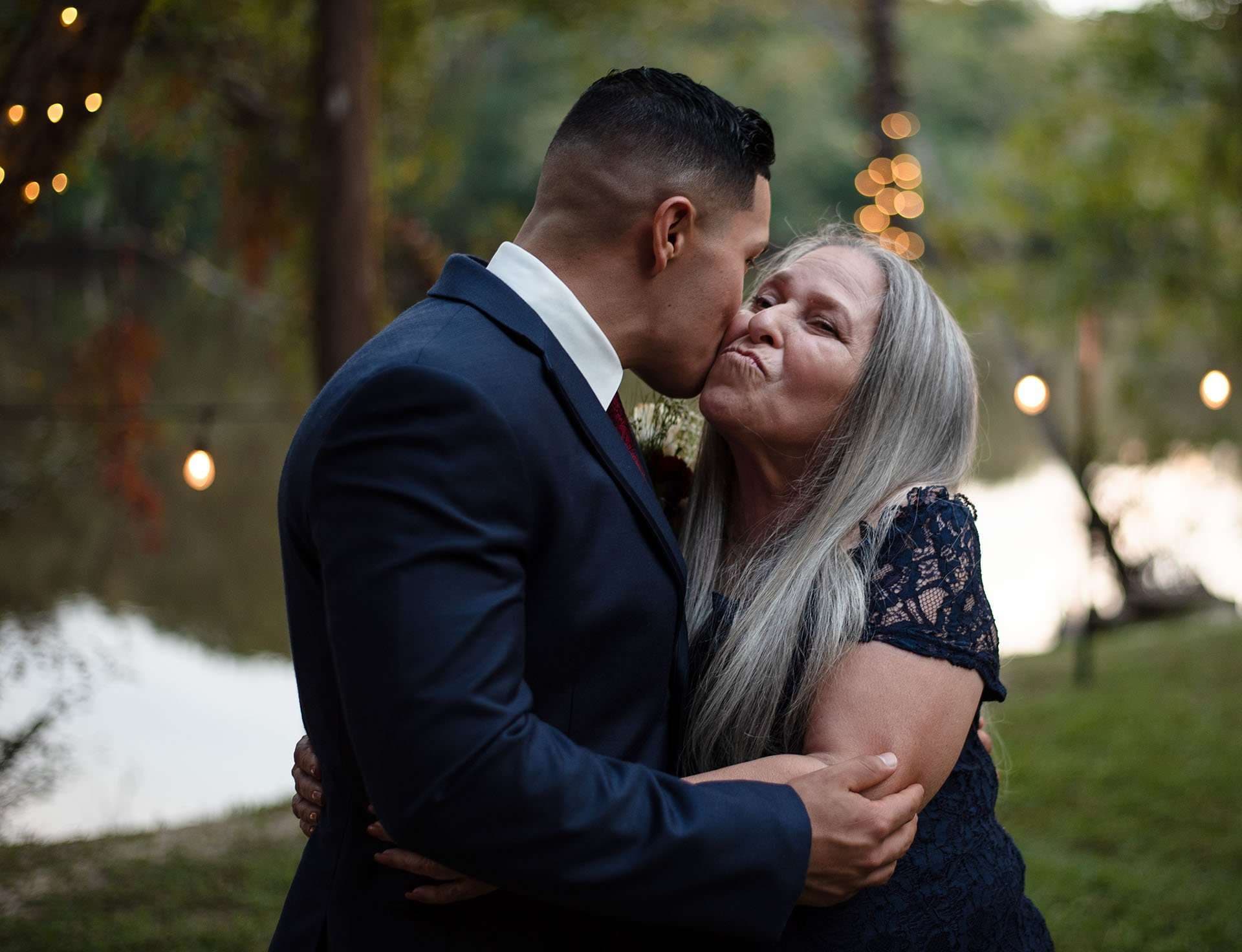 Man in a navy suit kisses the cheek of a gray-haired woman in a lace dress by a lake at dusk