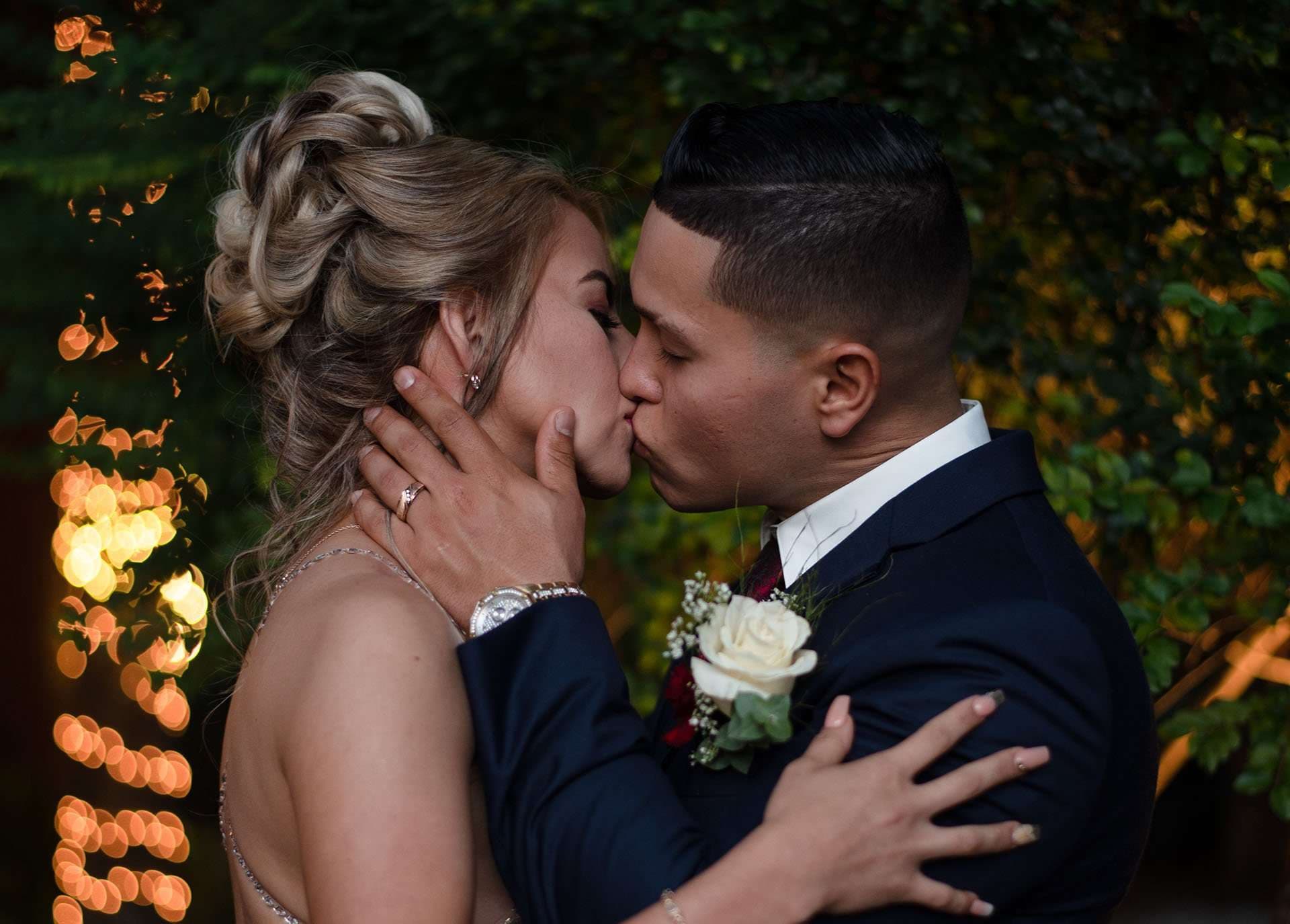Couple kissing outdoors at dusk, her hand on his cheek, he in a navy suit with a white rose boutonniere