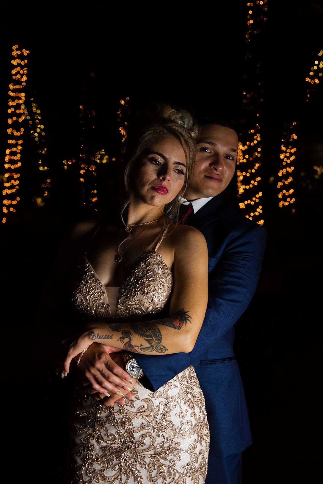 Couple embracing at night, man in a navy suit hugging a woman in a beaded gown from behind, string lights glowing