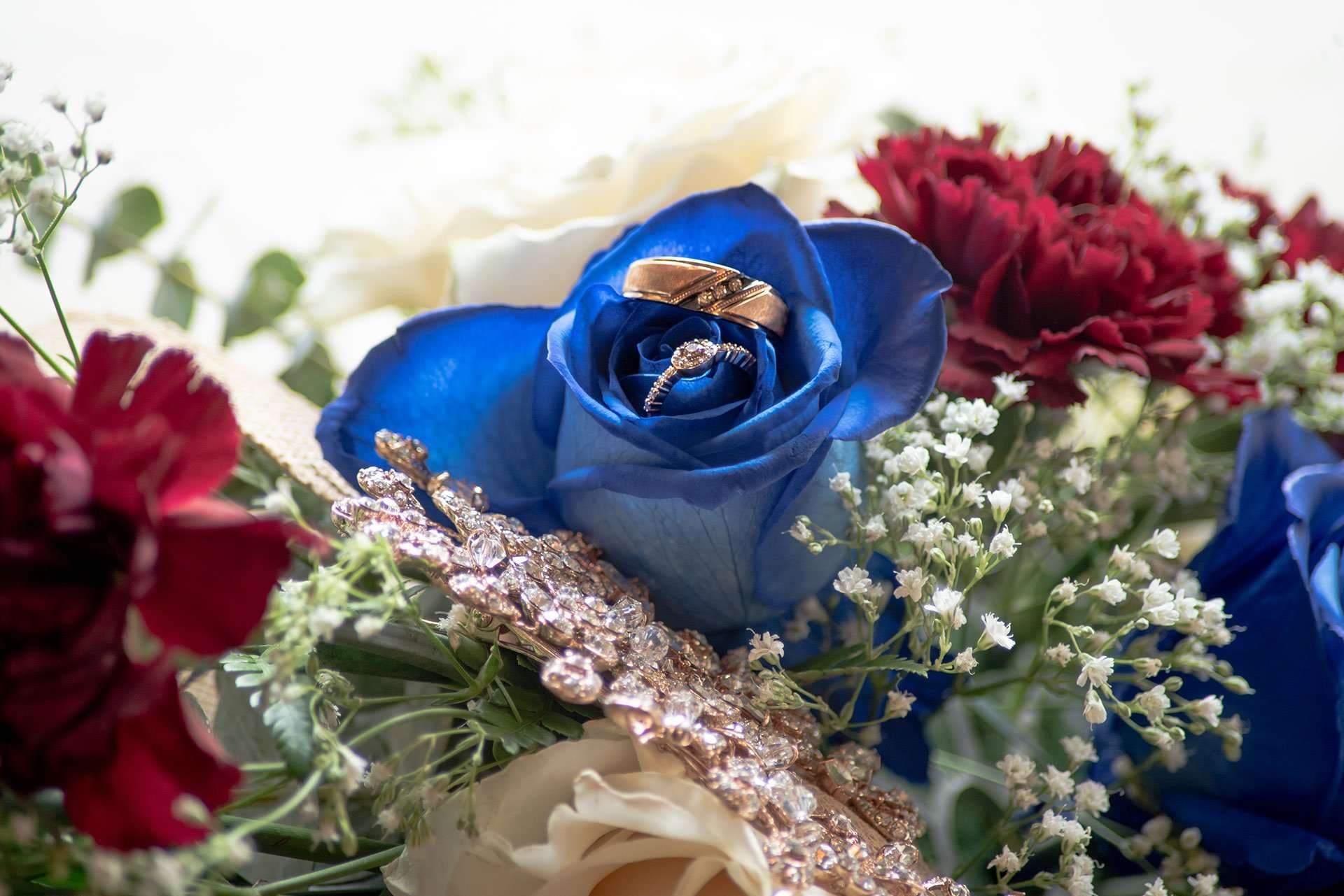Wedding rings resting inside a blue rose in a bouquet with red carnations, white baby's breath and a jeweled wrap