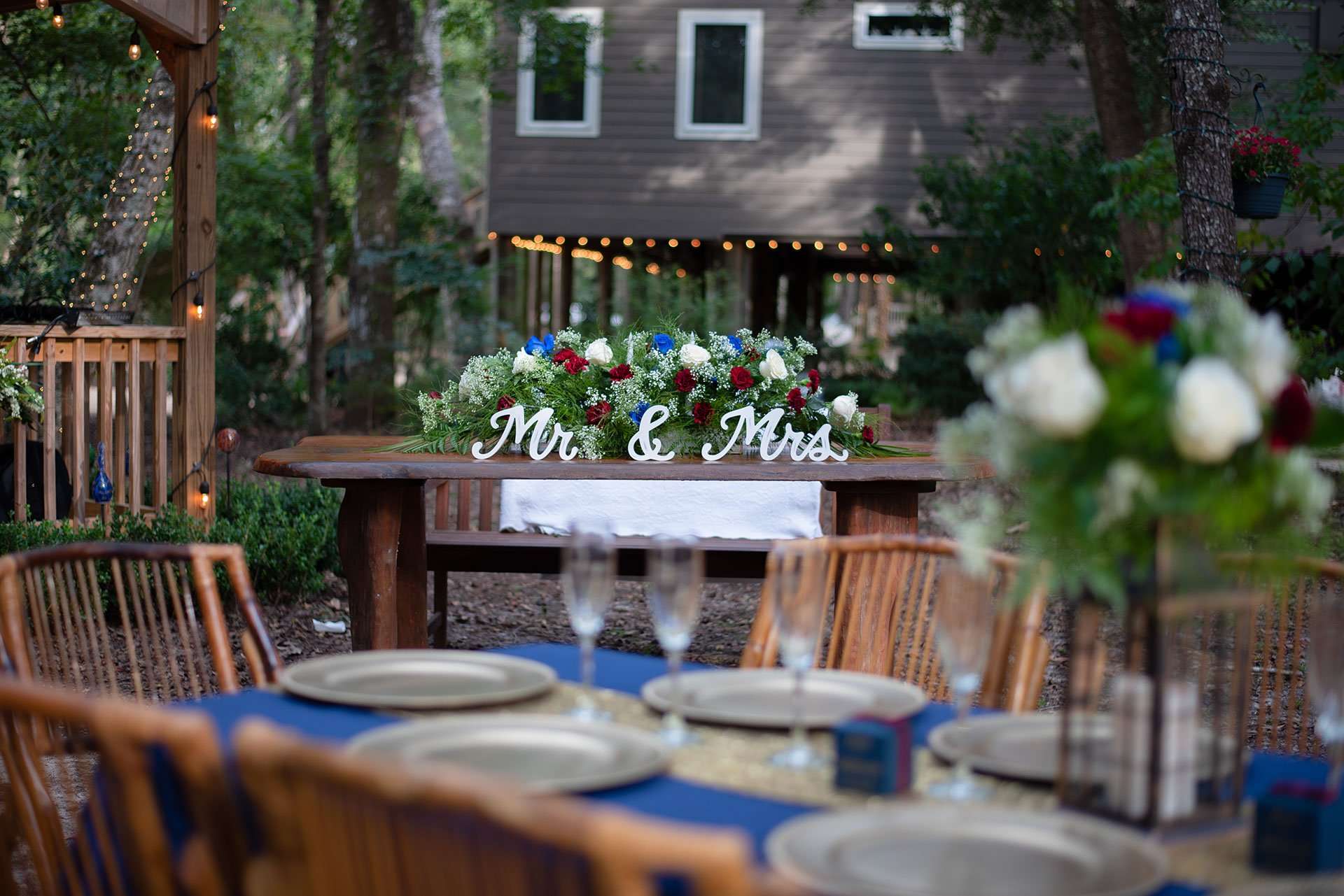 Outdoor wedding reception with a Mr & Mrs sign and red, white and blue floral arrangement on a wooden sweetheart table