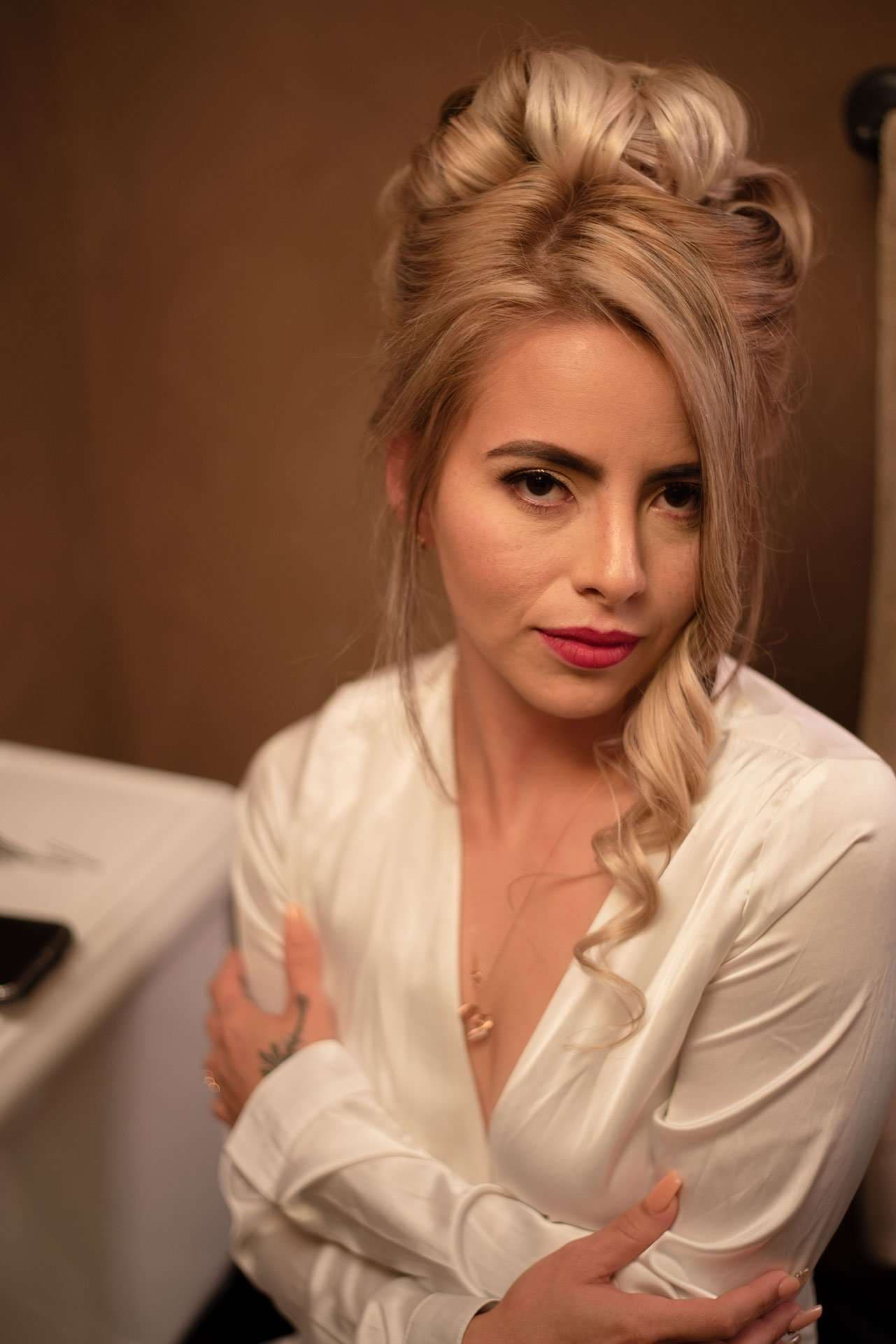Woman with blonde updo and red lipstick in a white satin robe, arms crossed, looking at the camera