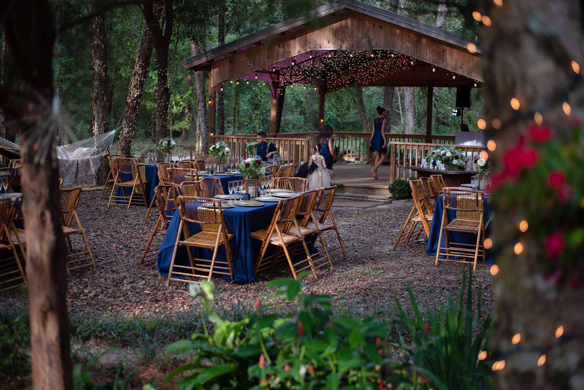 Outdoor wedding reception with blue-draped tables and bamboo chairs beneath string lights near a wooden pavilion in the woods