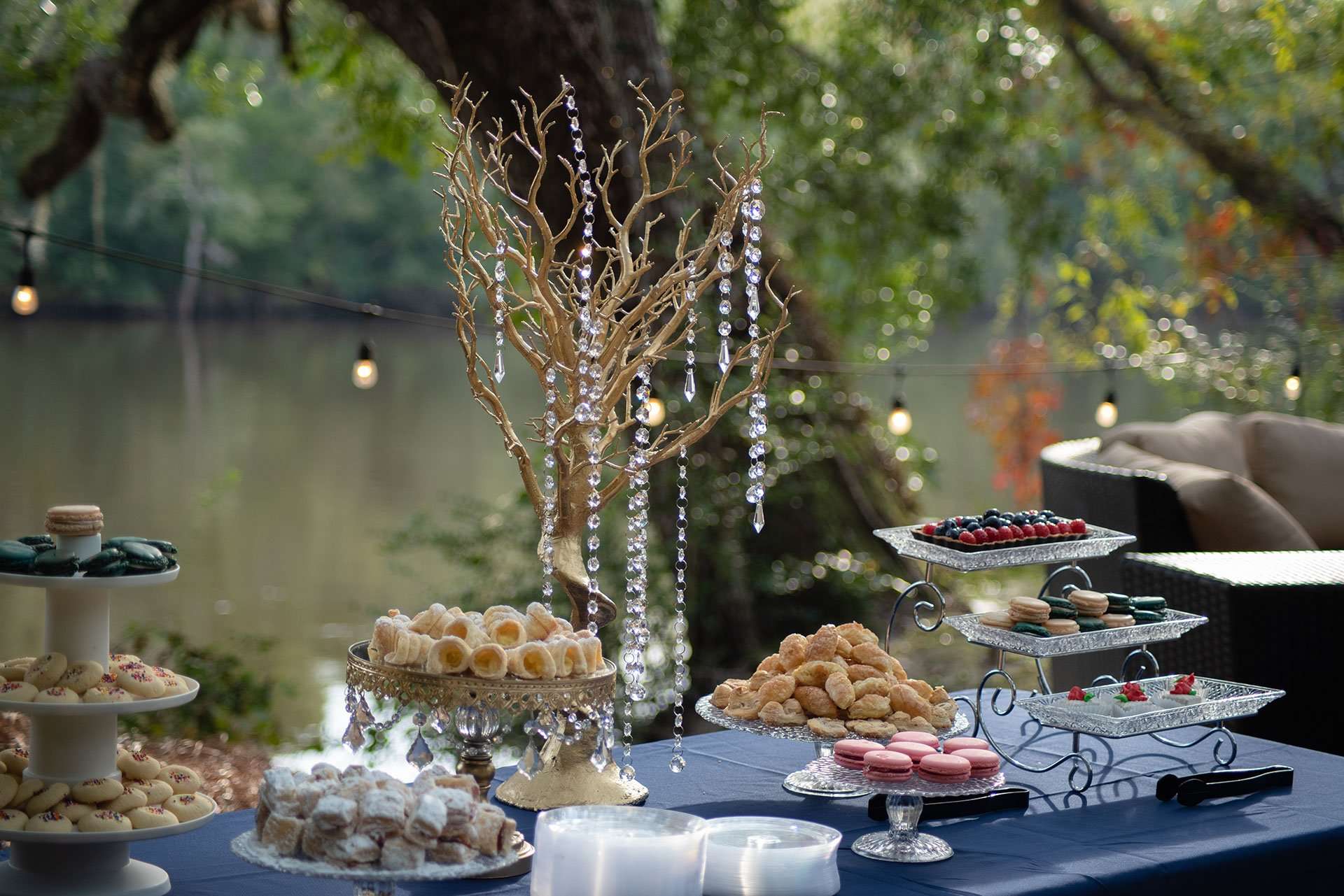 Dessert table with macarons, pastries and a crystal-draped gold branch centerpiece beside a calm lake at dusk