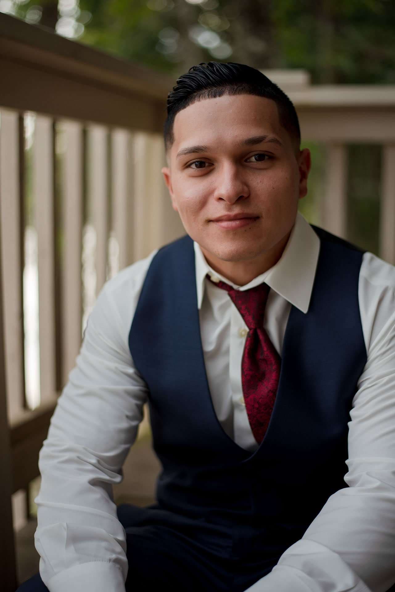Smiling groom in a navy vest, white shirt and burgundy tie seated beside a wooden railing