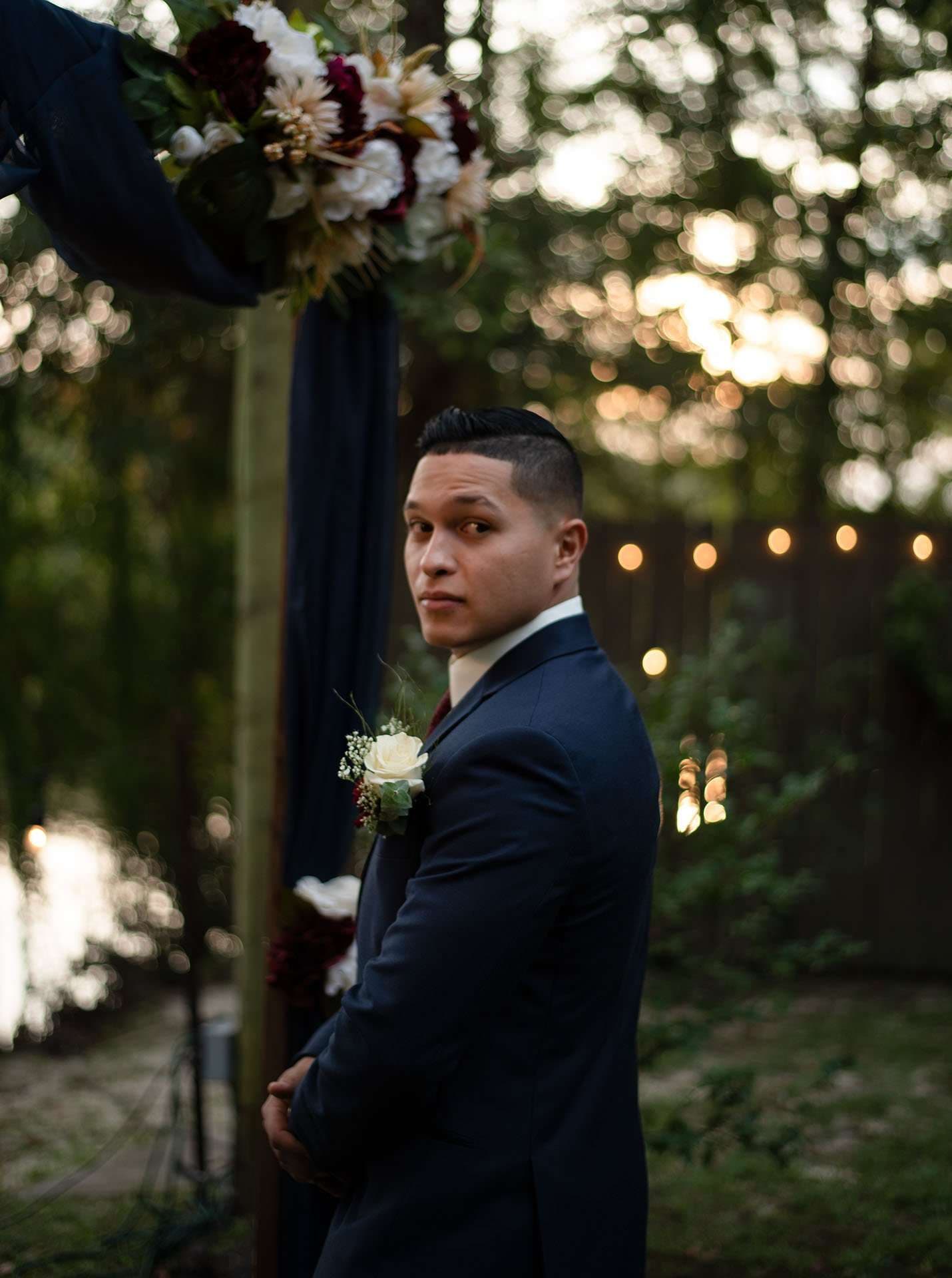 Groom in a navy suit and white rose boutonniere glances over his shoulder beside a floral arch with string lights