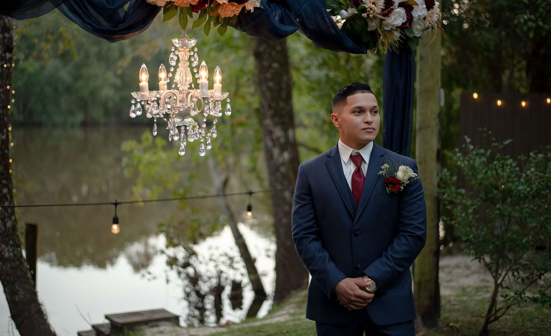 Groom in navy suit with red boutonniere waits under a crystal chandelier and draped fabric beside a lake