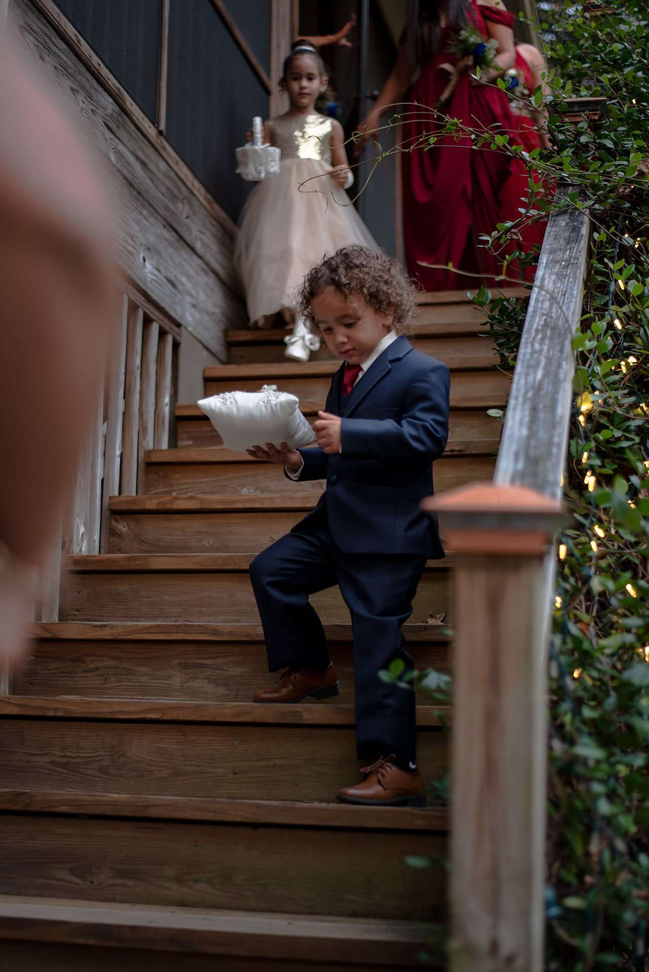 Ring bearer in a navy suit carries a white pillow down wooden stairs, followed by a flower girl in a tulle dress