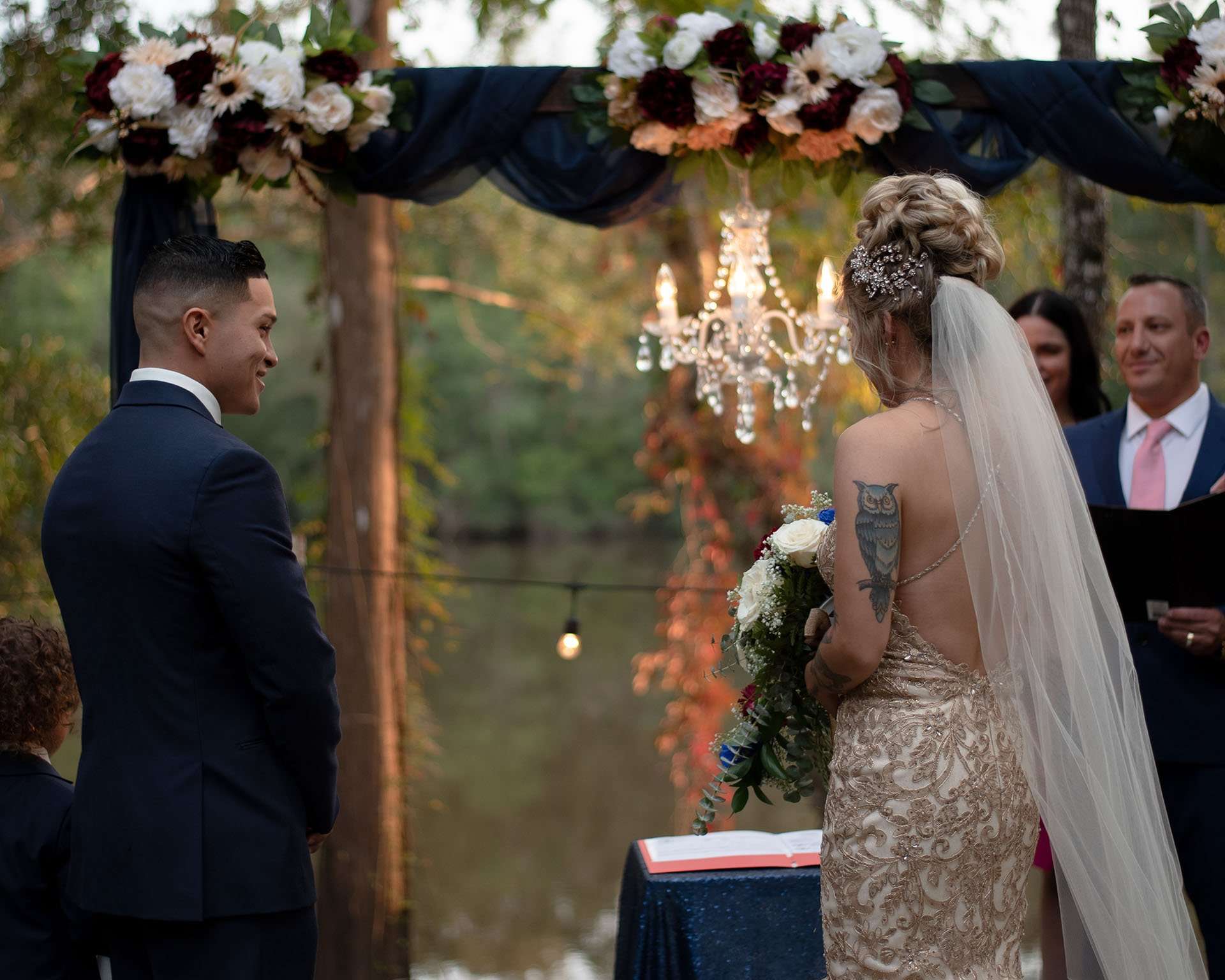 Groom in navy suit smiles at his bride in a gold lace gown and veil beneath a floral arch and chandelier