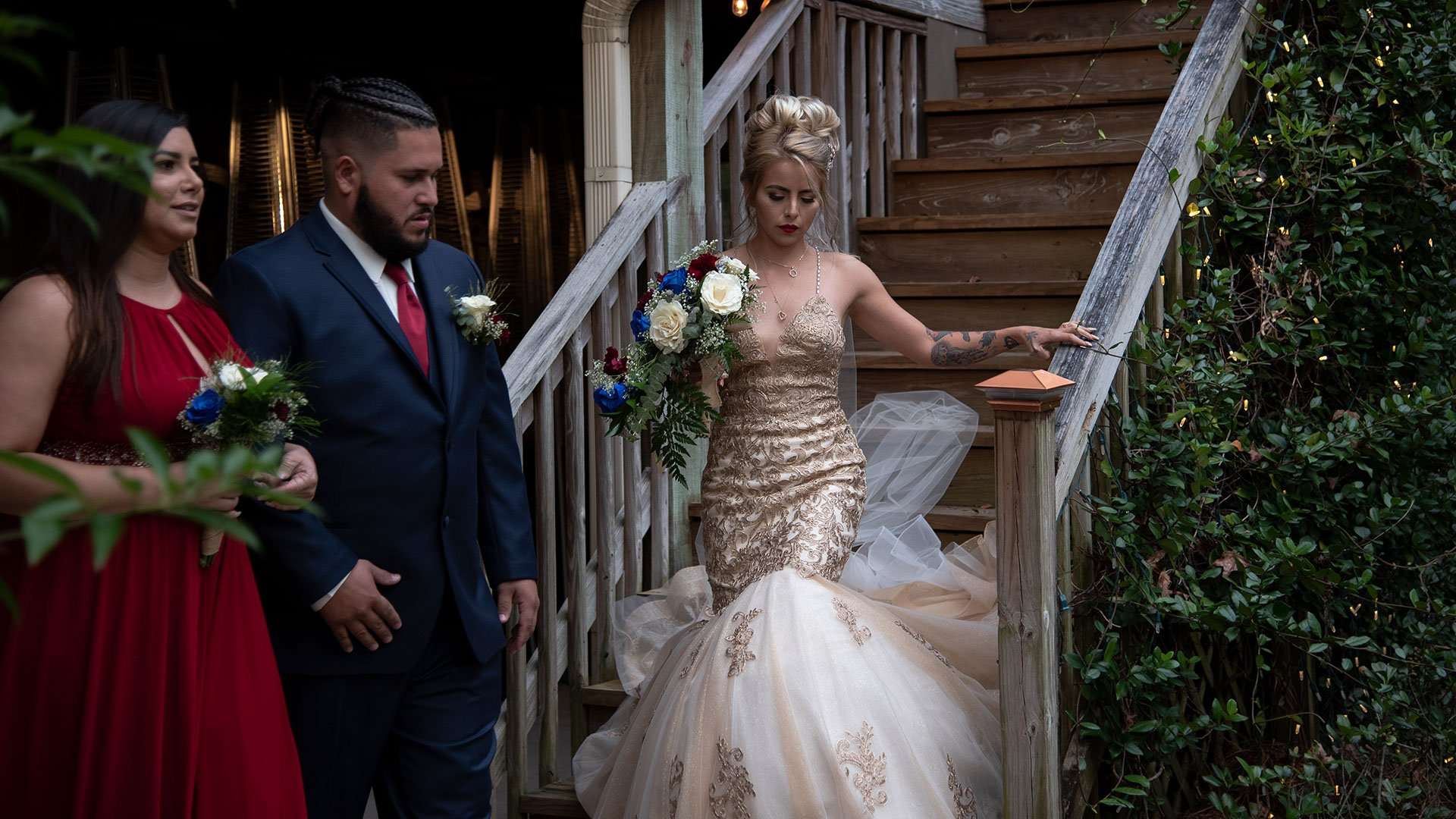 Bride in a gold sequined mermaid gown holding a bouquet walks down wooden stairs beside a groomsman and bridesmaid in red