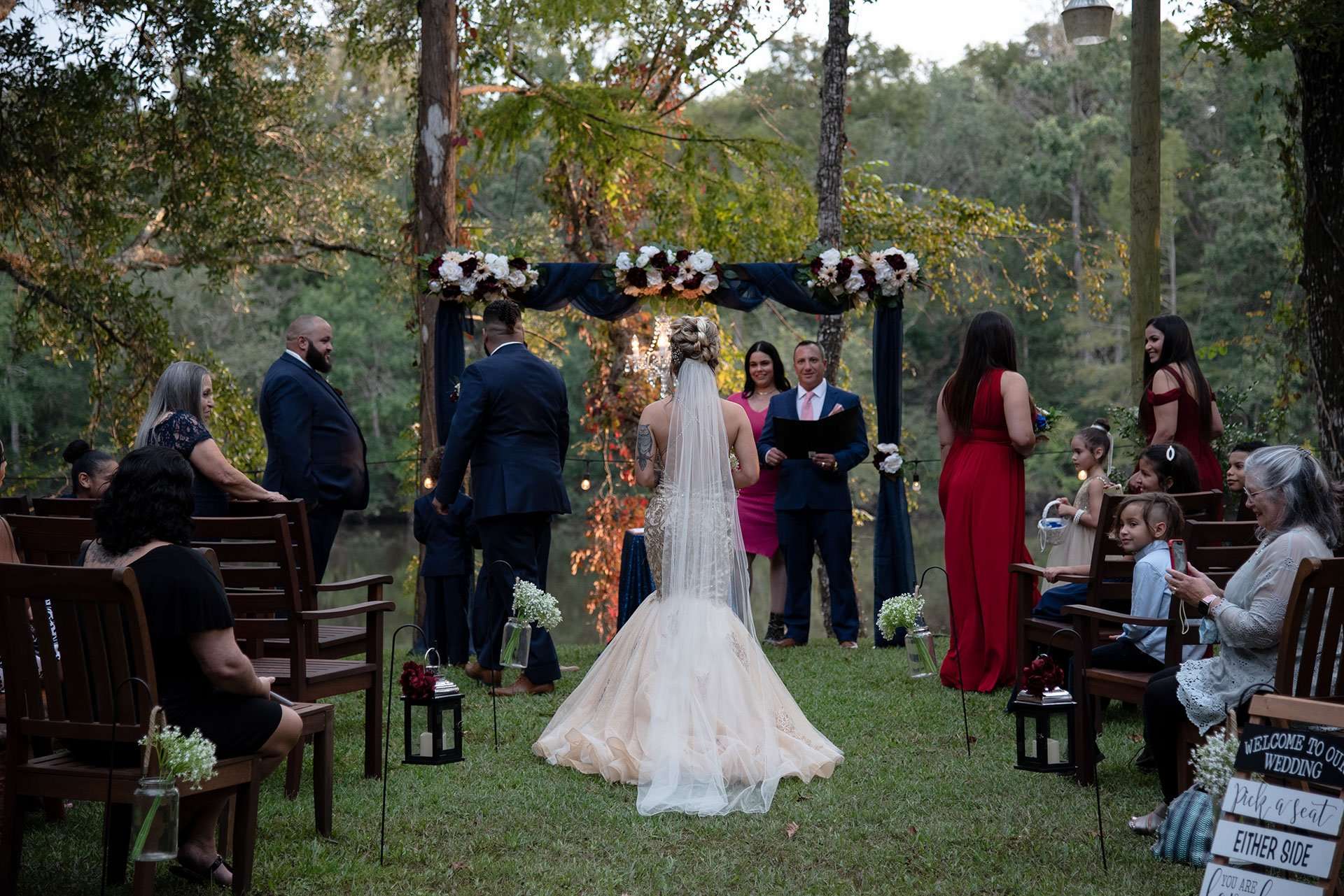 Bride in a long veil and blush gown faces the officiant under a floral arch as guests watch outdoors